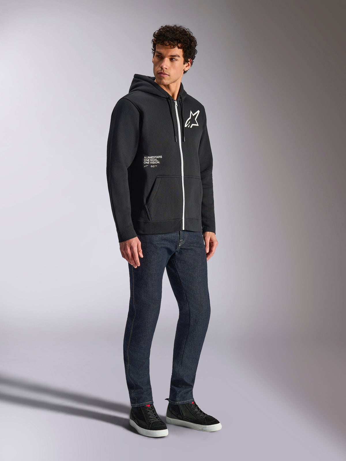 Camisola com capuz Alpinestars Transverse, sweatshirt preta com fecho de correr branco contrastante, apresenta um logótipo de estrela branco no peito e branding 'One Goal' no bolso, usada por um modelo com calças de ganga escuras e ténis pretos, streetwear casual inspirado no desporto motorizado