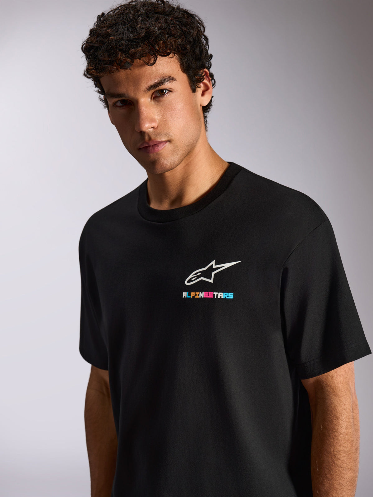 Um homem a usar a T-shirt Alpinestars Wild Oversized CSF, t-shirt preta de manga curta, com o logótipo Astars branco e grafismo multicolorido vibrante no peito, estilo casual motorsport streetwear