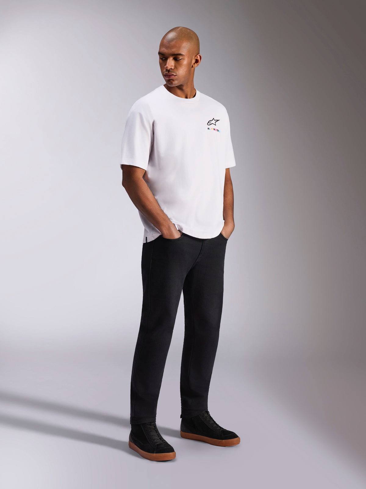 Alpinestars Wild Oversized CSF Tee, t-shirt de manga curta, branca, com um pequeno logótipo Astars multicolor no peito, usada por um modelo com calças pretas e sapatilhas pretas, streetwear casual inspirado no desporto motorizado