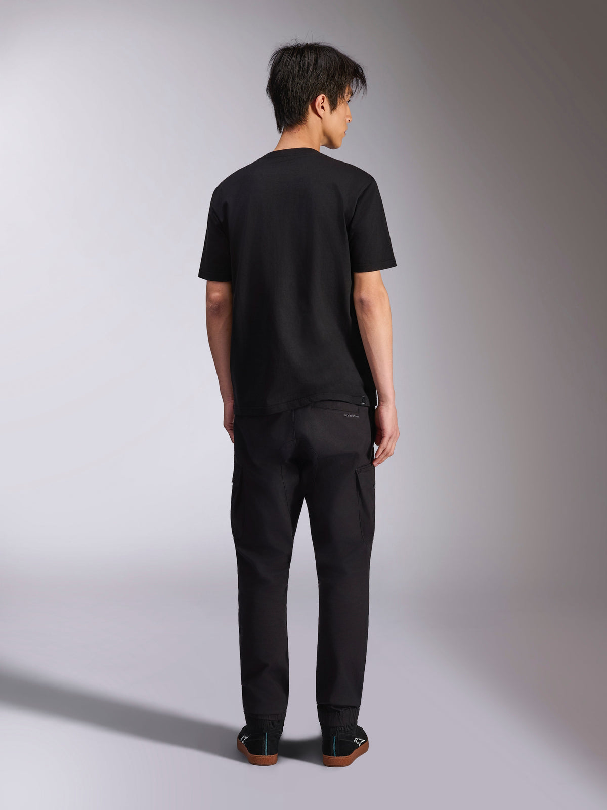 Alpinestars Noka CSF Tee, T-shirt de manga curta, preta, vista traseira de modelo a usar a t-shirt com calças pretas e sapatilhas, estilo casual motorsport streetwear