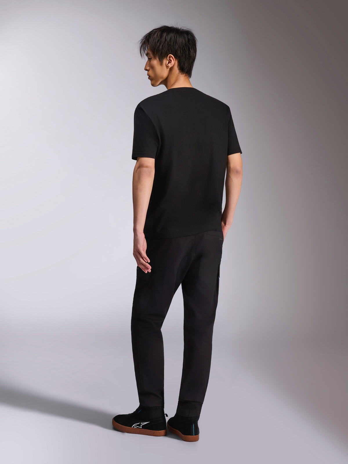 Alpinestars Noka CSF Tee, T-shirt de manga curta, preta, vista traseira num modelo, combinada com calças pretas e sapatilhas casuais, streetwear minimalista de motorsport
