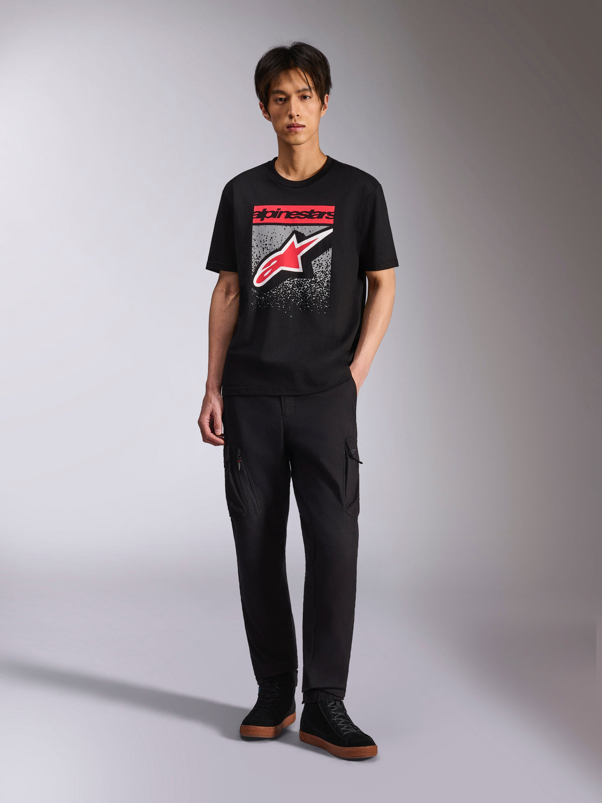 Alpinestars Noka CSF Tee, t-shirt de manga curta, preta, logótipo Astars frontal com gráfico de gradação MX em vermelho e branco, usada por um modelo com calças cargo pretas, Casual Motorsport Streetwear