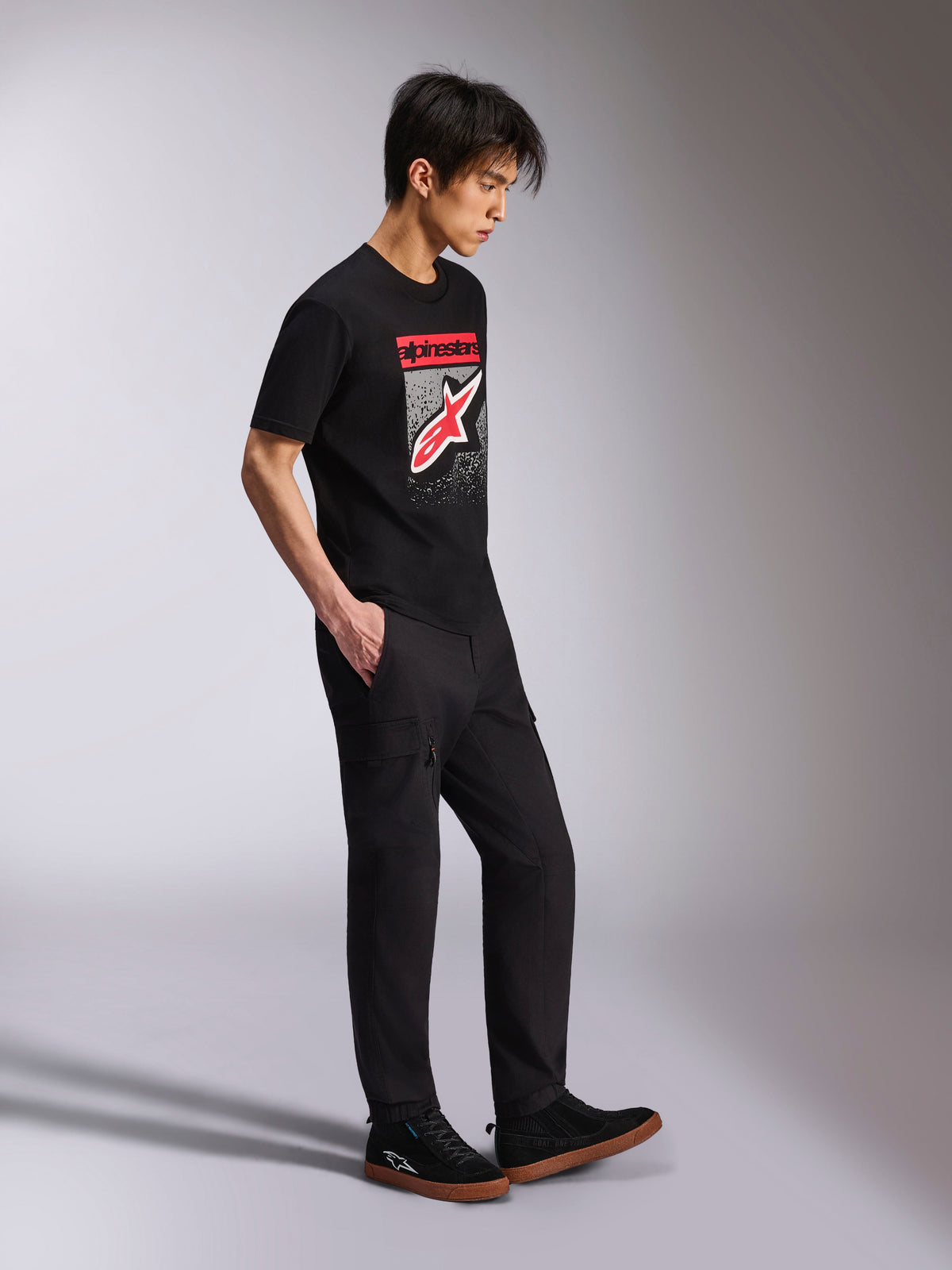 T-shirt Alpinestars Noka CSF, T-shirt de manga curta, preta, logótipo Astars frontal com gradação MX em vermelho e branco, usada com calças cargo pretas e sapatilhas, streetwear casual inspirado no desporto motorizado