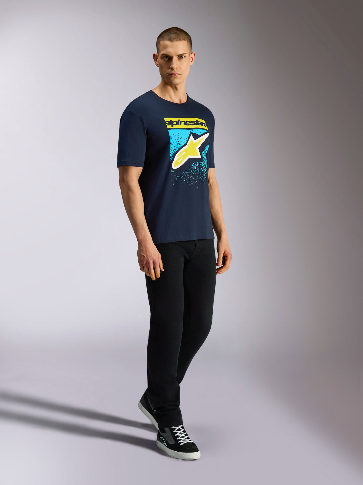 Alpinestars Noka CSF Tee, t-shirt de manga curta, azul-marinho, logótipo Astars amarelo com gráfico de gradação MX azul, modelo masculino a usar t-shirt azul-marinho com calças pretas e sapatilhas, streetwear casual de desportos motorizados