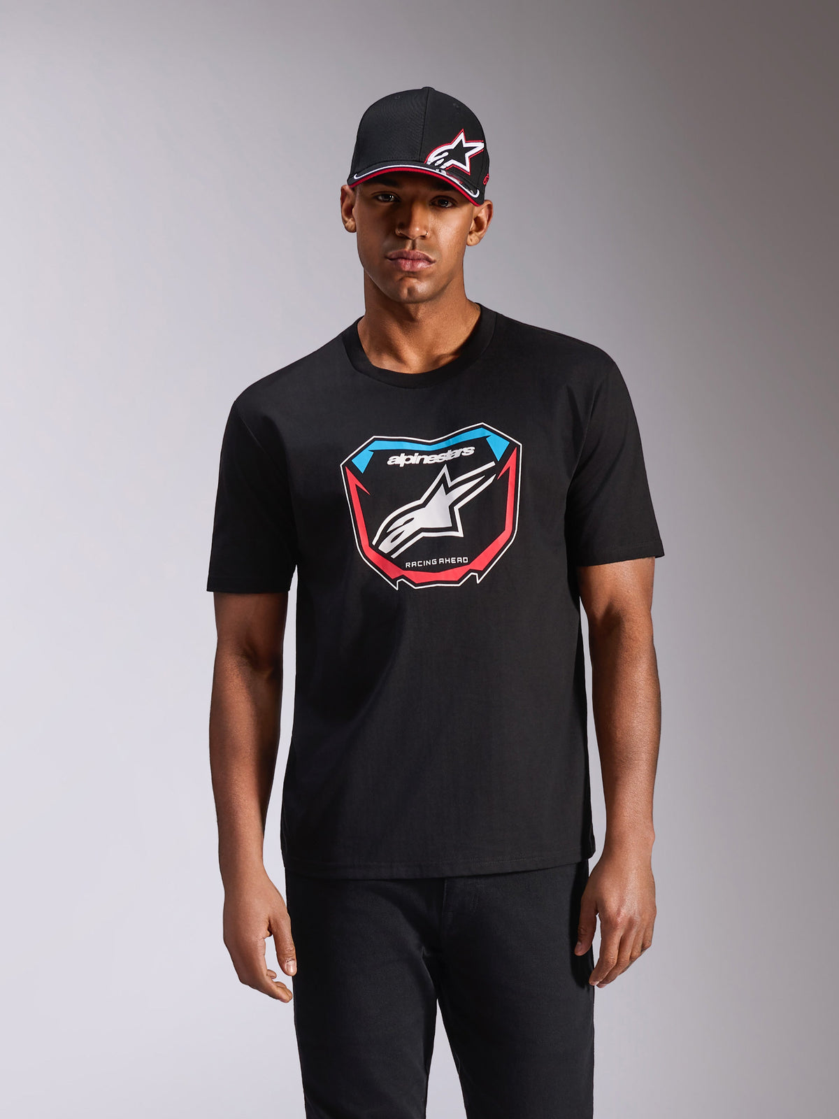 Alpinestars Plate CSF Tee, t-shirt de manga curta, preta, com um gráfico frontal de placa MX, logótipo branco e detalhes em vermelho e azul, usada por um modelo com boné preto a condizer, streetwear casual de desportos motorizados