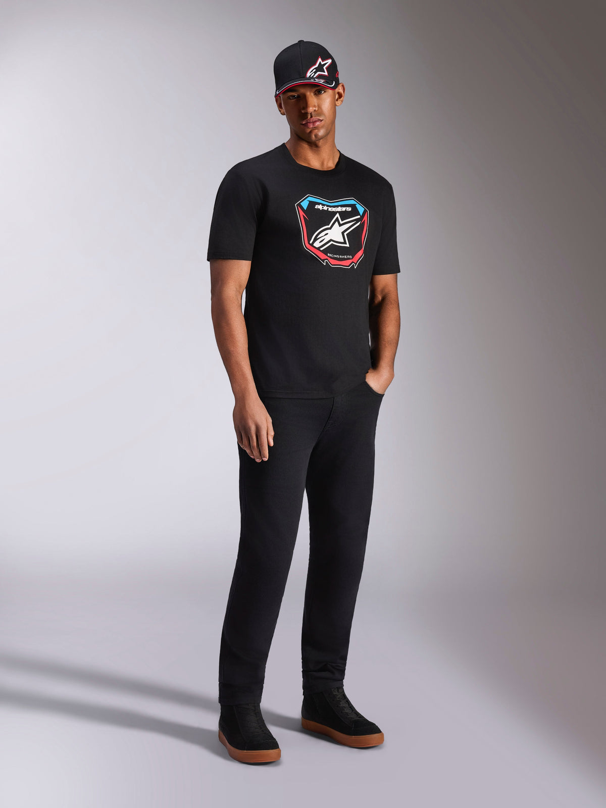 Alpinestars Plate CSF Tee, t-shirt de manga curta, preta, gráfico frontal colorido de placa MX com logótipo de estrela, usada por modelo com boné preto com logótipo e calças, streetwear casual inspirado no desporto motorizado