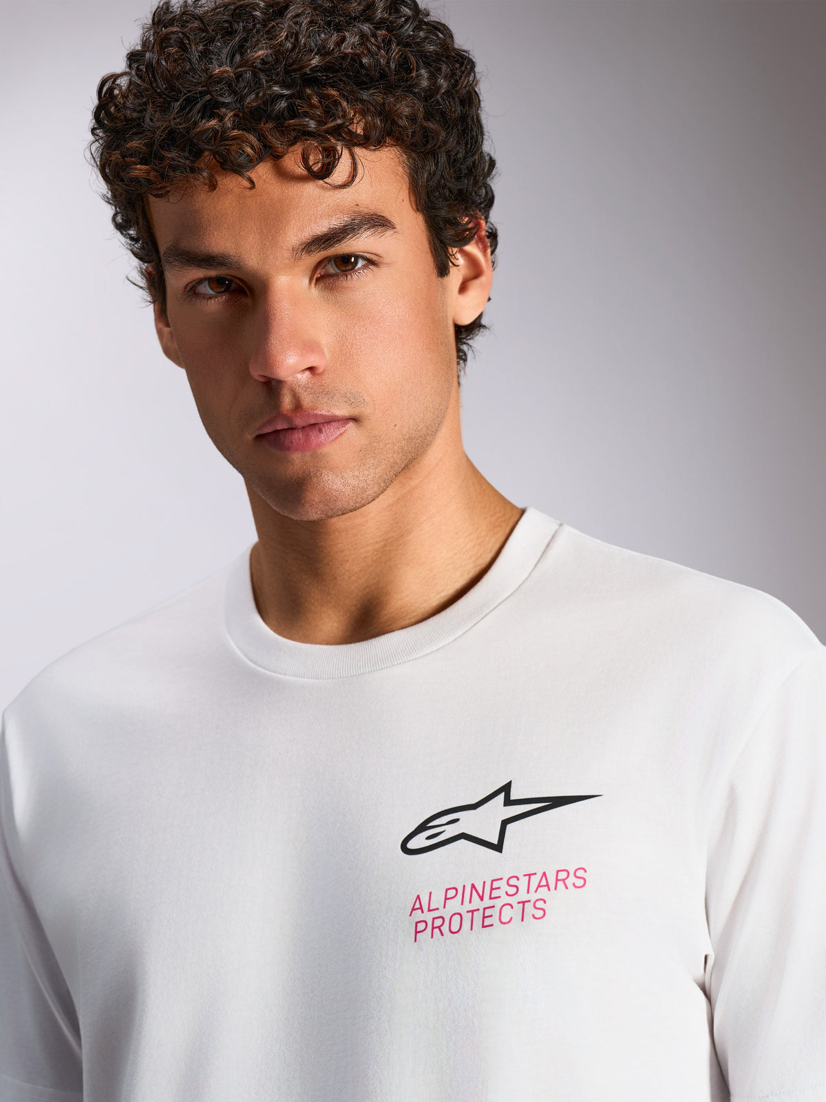 T-shirt Alpinestars Pop Helmet CSF, t-shirt de manga curta branca, logótipo Astars preto e texto cor-de-rosa 'ALPINESTARS PROTECTS' no peito, streetwear casual de desportos motorizados