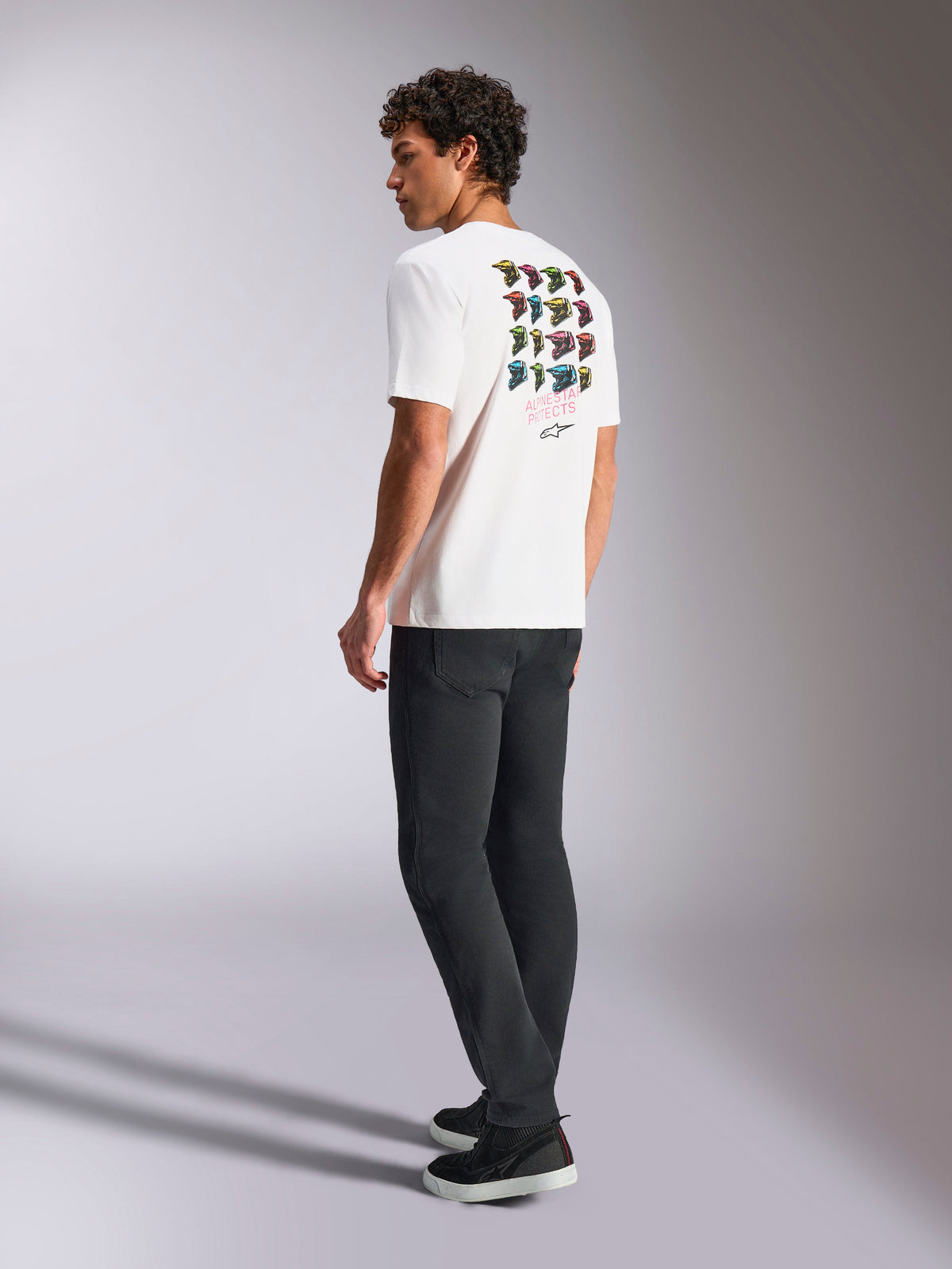 Alpinestars Pop Helmet CSF Tee, t-shirt de manga curta branca, vista traseira no modelo a exibir a arte gráfica colorida do capacete M10, combinada com calças escuras e sapatilhas pretas, streetwear casual inspirado no desporto motorizado