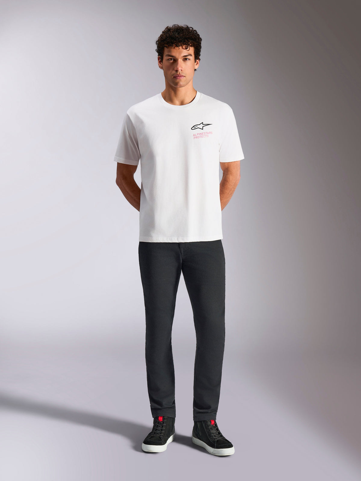T-shirt Alpinestars Pop Helmet CSF, t-shirt de manga curta, branca, vestida por um modelo com calças pretas e sapatilhas de cano alto pretas, logótipo Astars frontal com texto rosa 'Alpinestars Protects', streetwear casual de motorsport