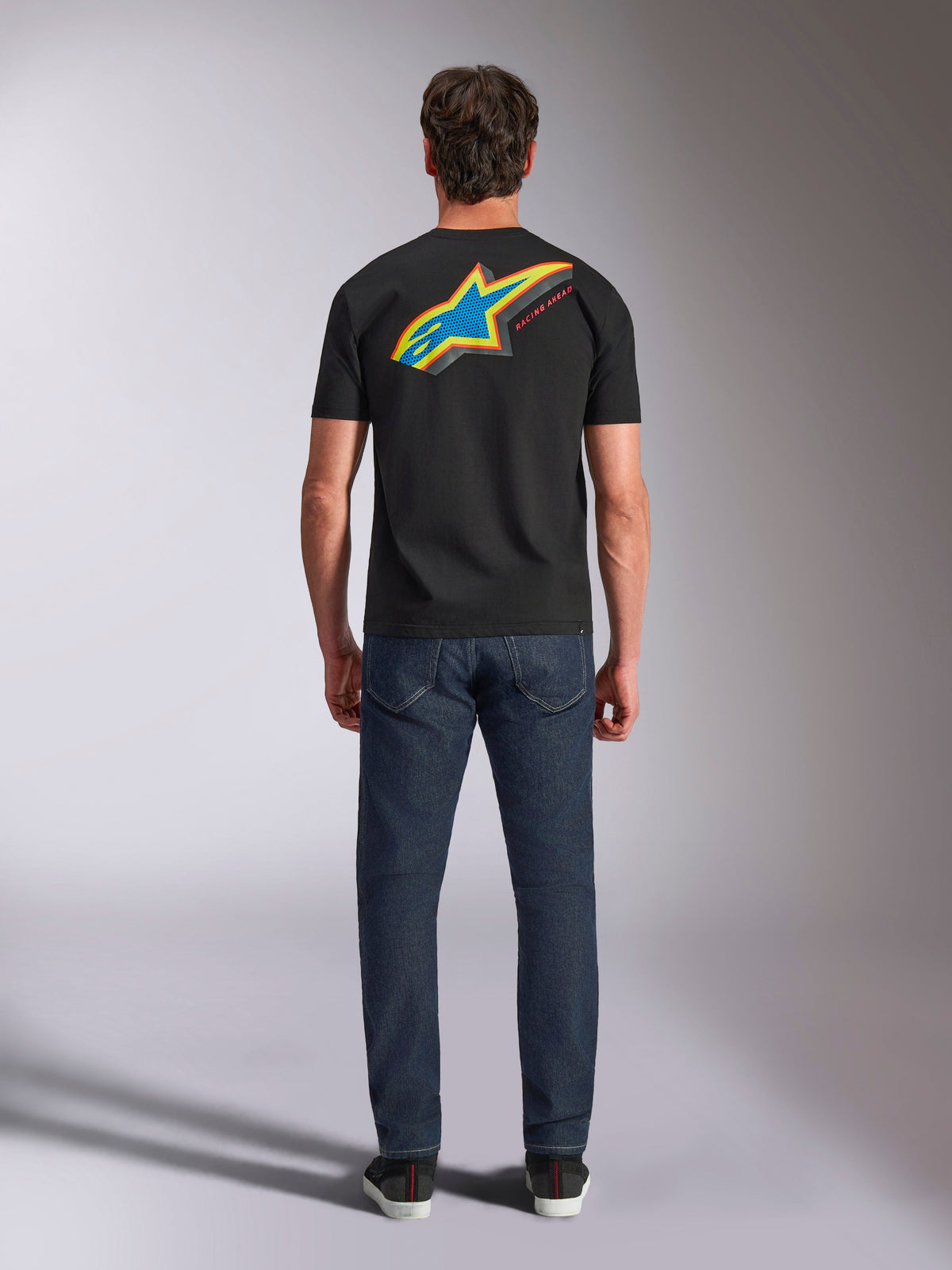 Alpinestars Angler CSF Tee, t-shirt de manga curta, preta, vista traseira do modelo a usar a t-shirt com logótipo de estrela Astars grande, vibrante e multicolor e texto 'RACING AHEAD', combinada com calças de ganga azul-escuro, streetwear casual de motorsport