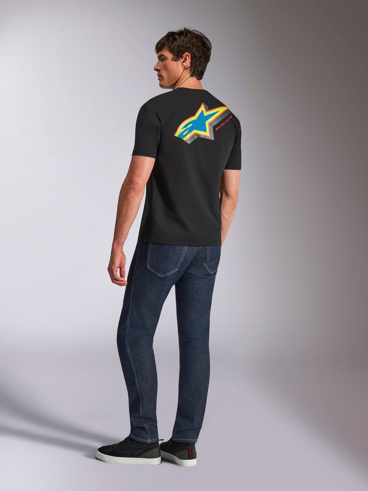 Alpinestars Angler CSF Tee, t-shirt de manga curta, preta, com um grande logótipo Astars vibrante e multicolorido nas costas com detalhes perfurados a azul, amarelo e vermelho, usada com calças de ganga azul-escuro, streetwear casual de desportos motorizados