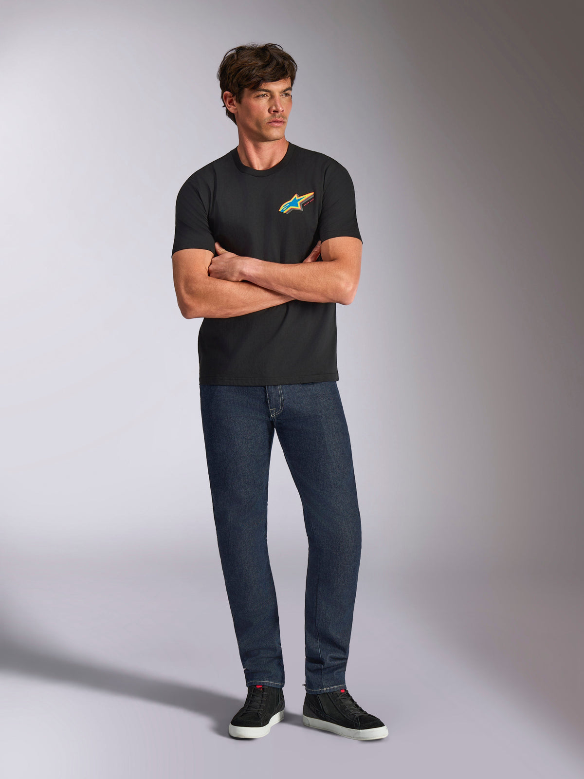 Alpinestars Angler CSF Tee, t-shirt de manga curta, preta, logótipo Astars vibrante e multicolor no peito, usada por modelo com os braços cruzados, calças de ganga escura e ténis pretos, streetwear casual de motorsport