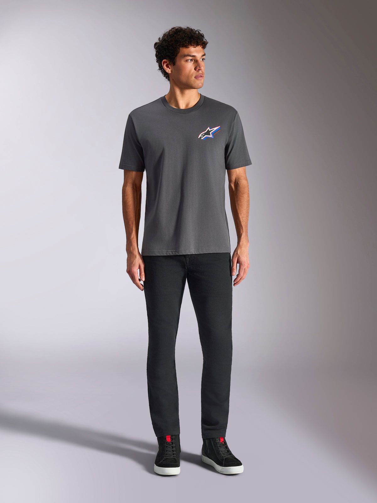 Alpinestars Angler CSF Tee, t-shirt de manga curta, cor Charcoal, com um logótipo Astars vibrante e multicolor no peito, vestida por um modelo com calças pretas e sapatilhas, Casual Motorsport Streetwear