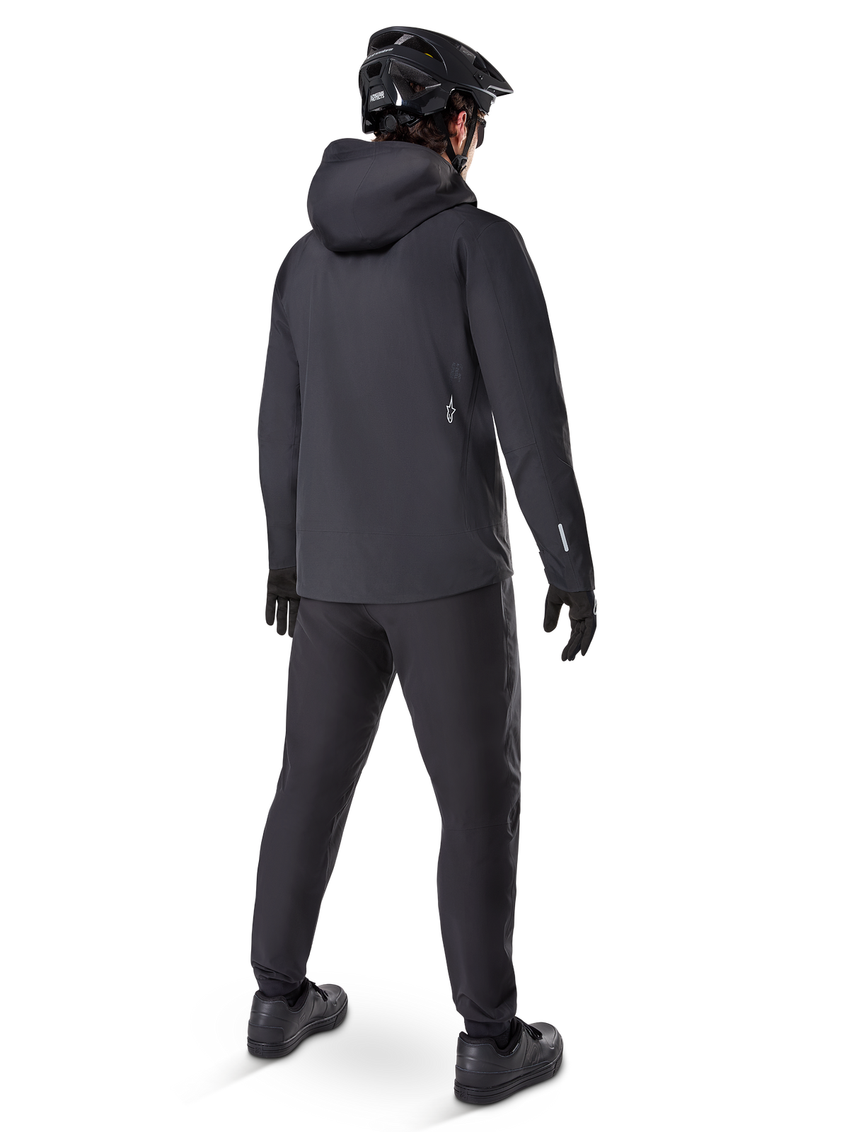 Casaco Impermeável Alpinestars A-Dura Elite, preto, vista traseira que mostra o capuz sobre o capacete e a bainha traseira mais longa, combinado com calças de BTT, luvas e capacete pretos a condizer para uma proteção completa contra as intempéries nos trilhos.