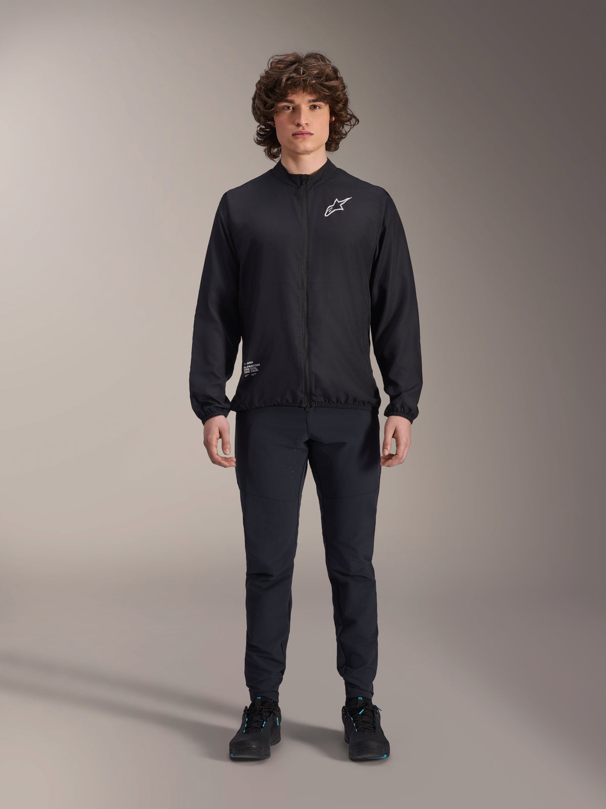 Modelo a usar o Casaco Corta-Vento Alpinestars A-Aria em preto, com fecho de correr frontal completo, gola alta e logótipo Astars branco no peito, combinado com calças técnicas pretas e sapatos pretos, vestuário de ciclismo de performance para BTT e uso ao ar livre.