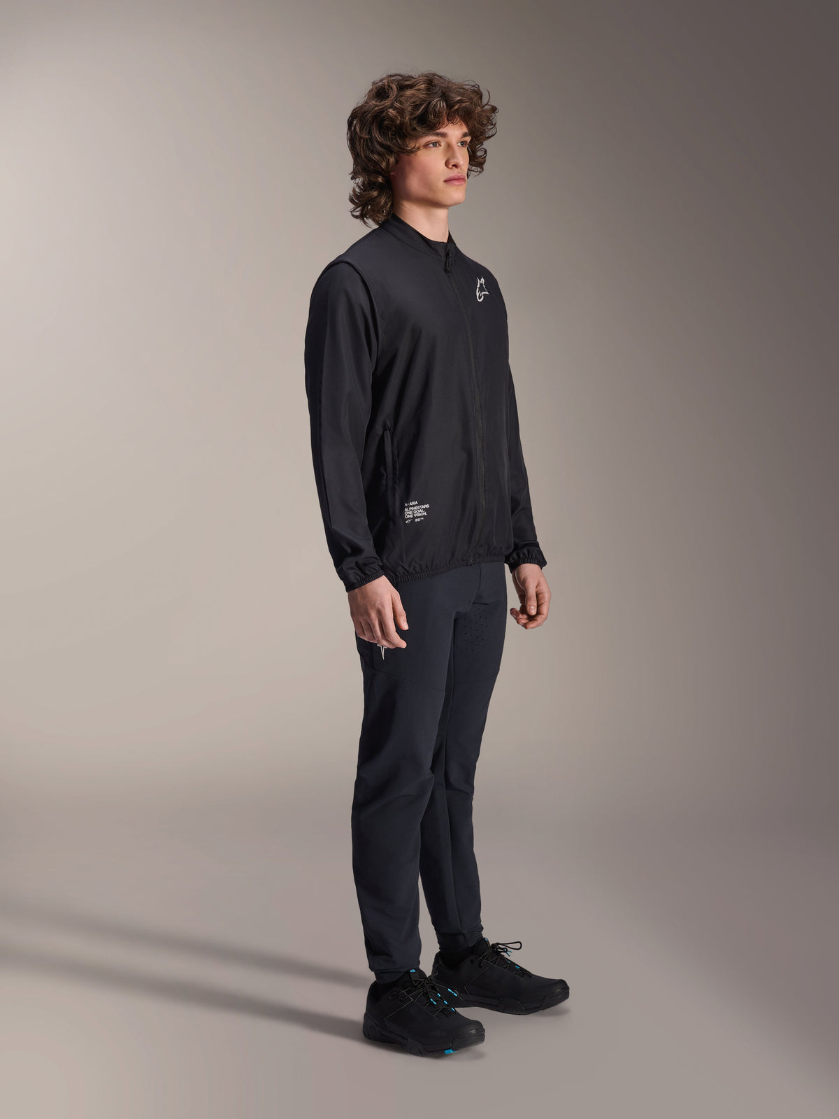 Casaco Corta-Vento Alpinestars A-Aria, preto, casaco de ciclismo leve e resistente ao vento vestido por um modelo com cabelo encaracolado, com um logótipo Astars branco no peito e marca na parte inferior direita, combinado com calças e sapatos pretos, corte de performance para BTT e gravel