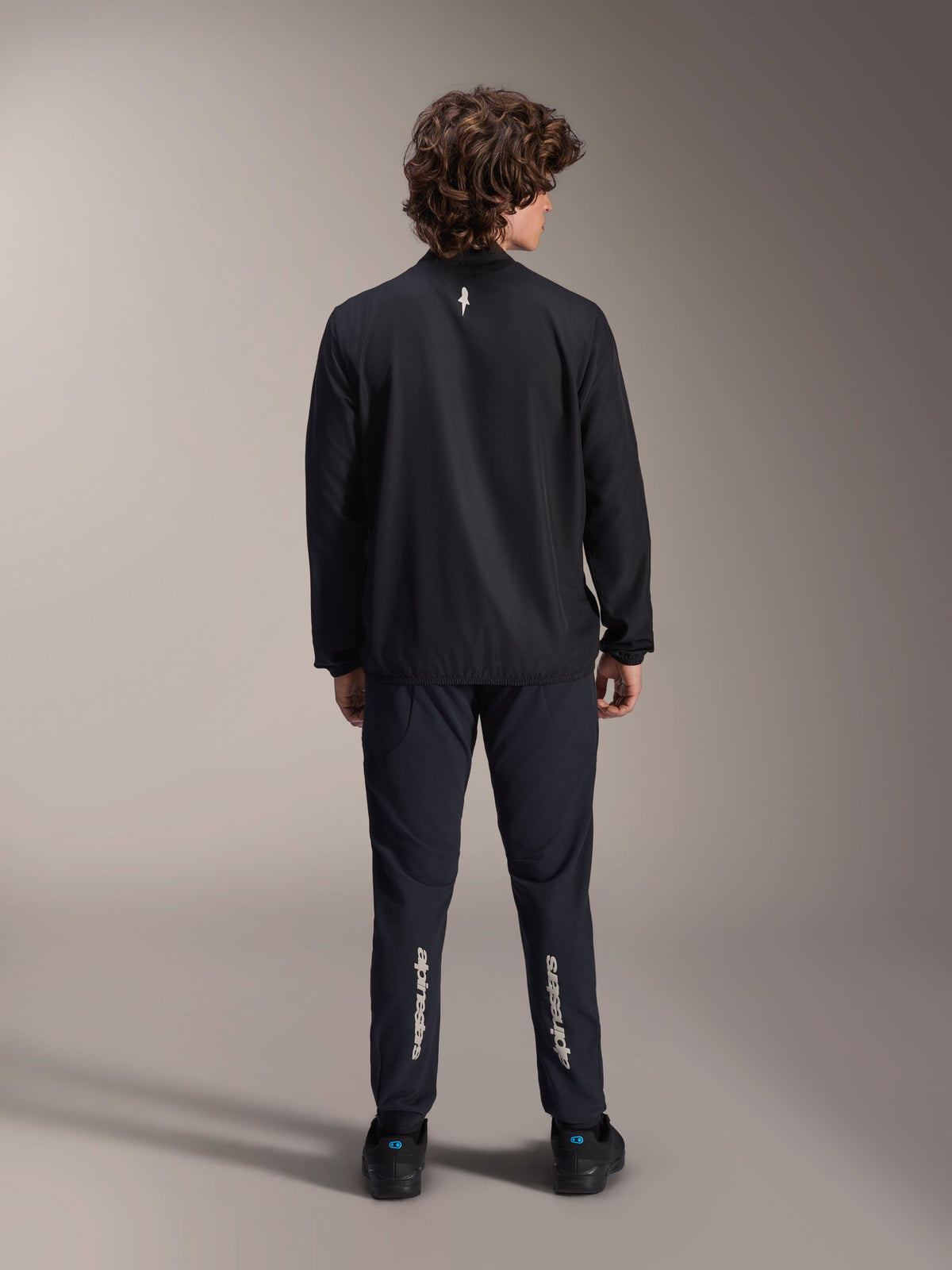 Casaco Corta-Vento Alpinestars A-Aria, preto, vista traseira, casaco de ciclismo leve e resistente ao vento com um pequeno logótipo branco na parte superior das costas, usado com calças pretas a condizer com a marca Alpinestars vertical na zona dos gémeos, equipamento de performance para mountain bike