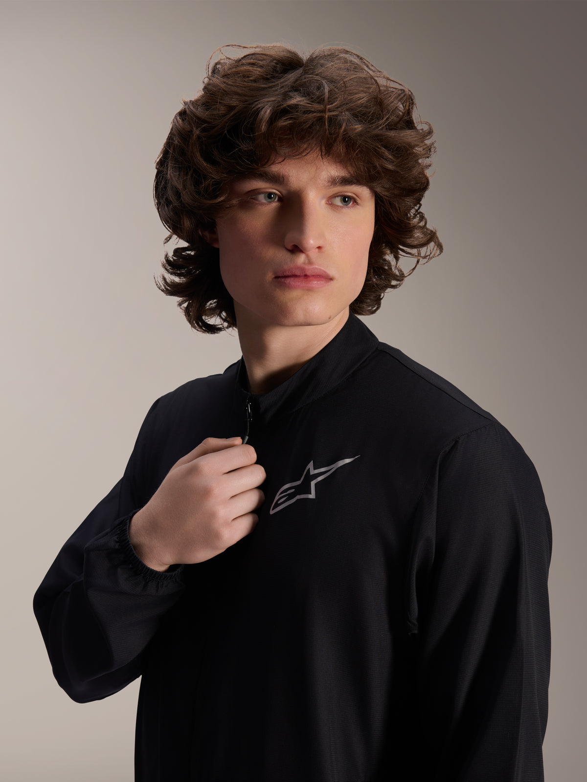 Casaco Corta-Vento Alpinestars A-Aria, casaco de ciclismo preto, vestido por um modelo masculino, design de gola alta com um logótipo Astars prateado no peito, equipamento de MTB de performance leve e resistente ao vento
