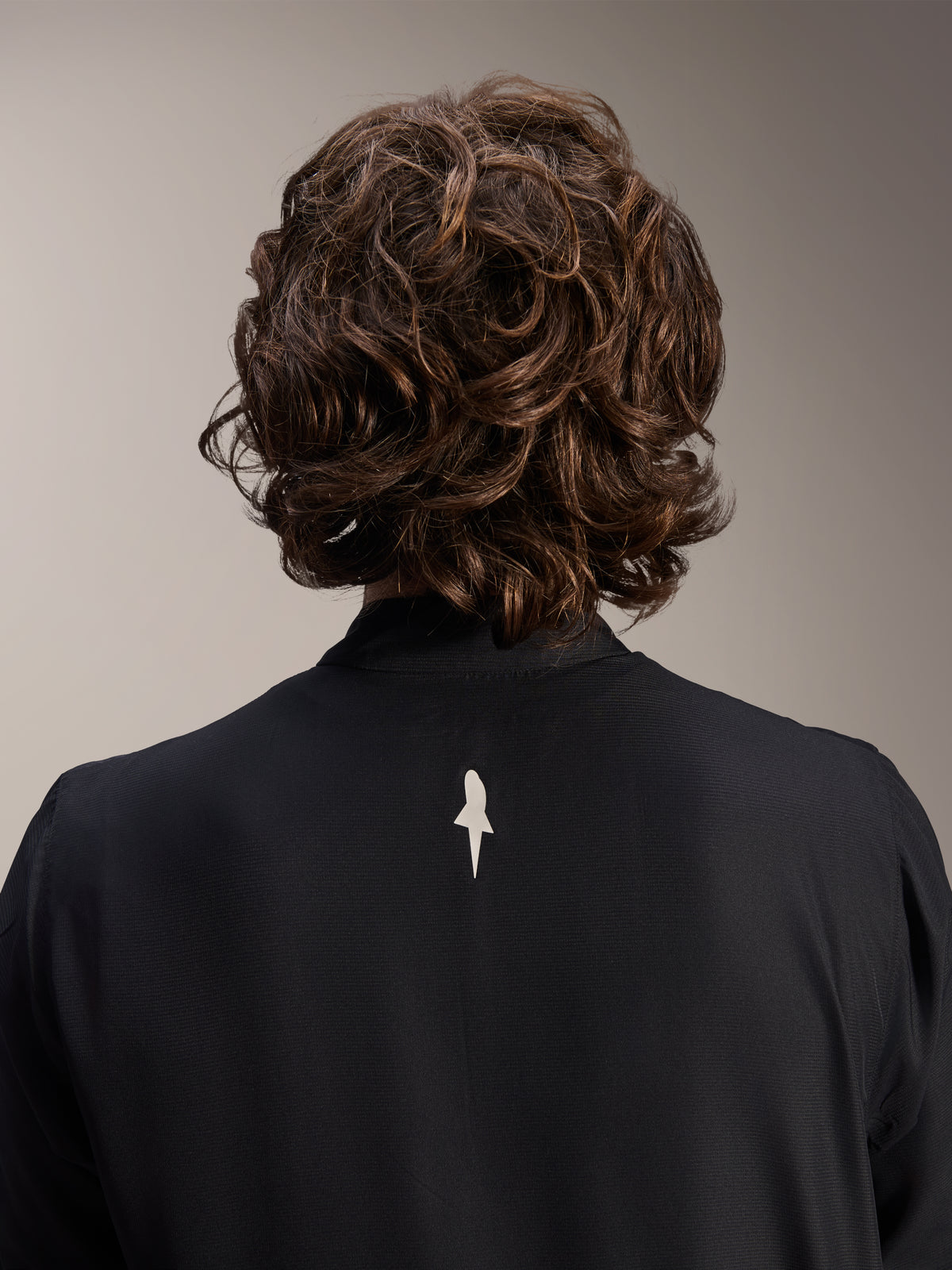 Casaco Corta-Vento Alpinestars A-Aria, preto, vista traseira em pormenor da zona superior dos ombros e gola, com um pequeno logótipo branco no centro das costas, usado por modelo com cabelo castanho ondulado, equipamento de ciclismo de performance minimalista