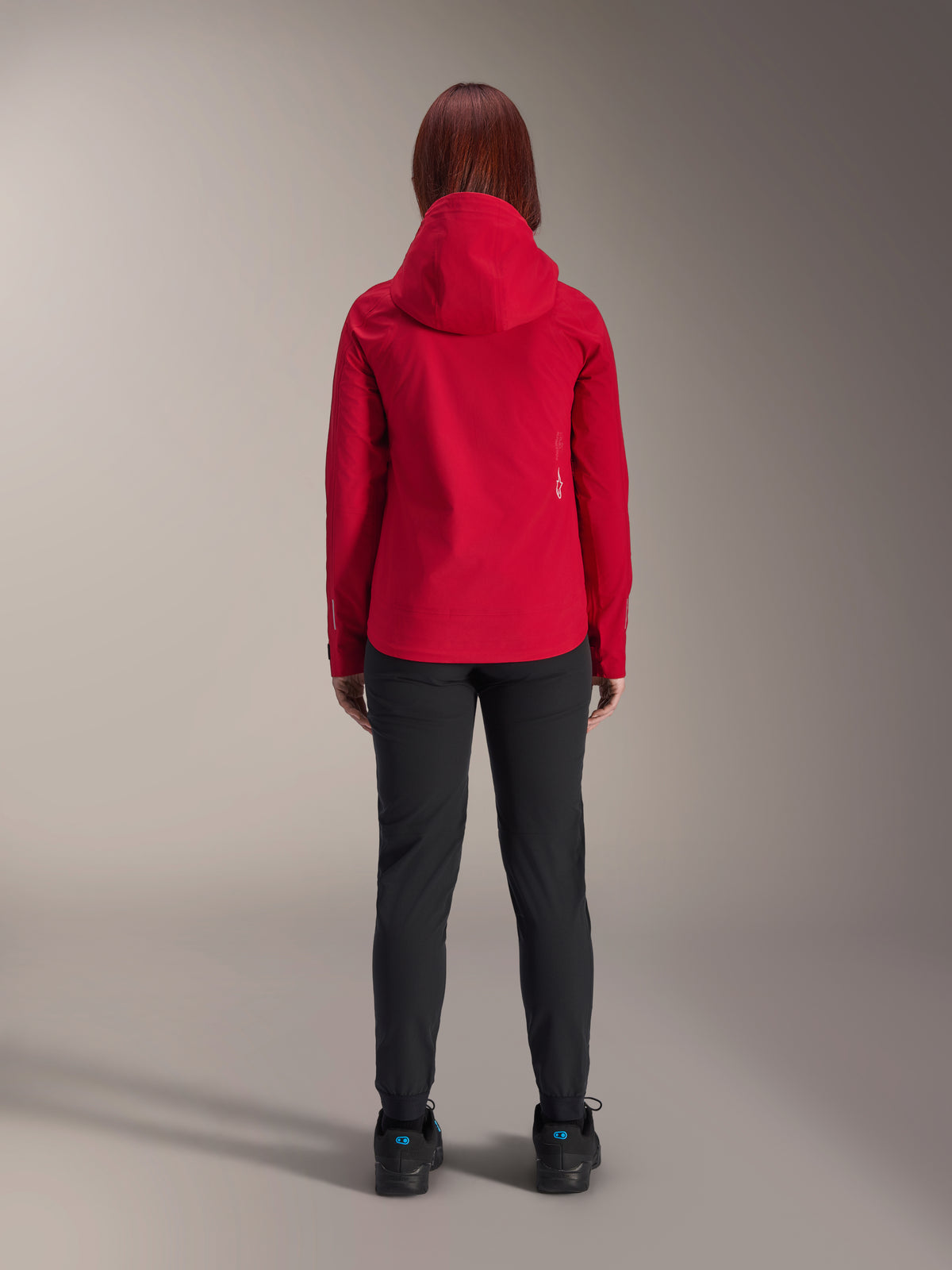 Vista traseira de uma mulher a usar o casaco impermeável Stella A-Dura Elite da Alpinestars em Vermelho Fluo, combinado com calças e sapatos pretos, com um capuz protetor e bainha traseira mais longa para BTT.