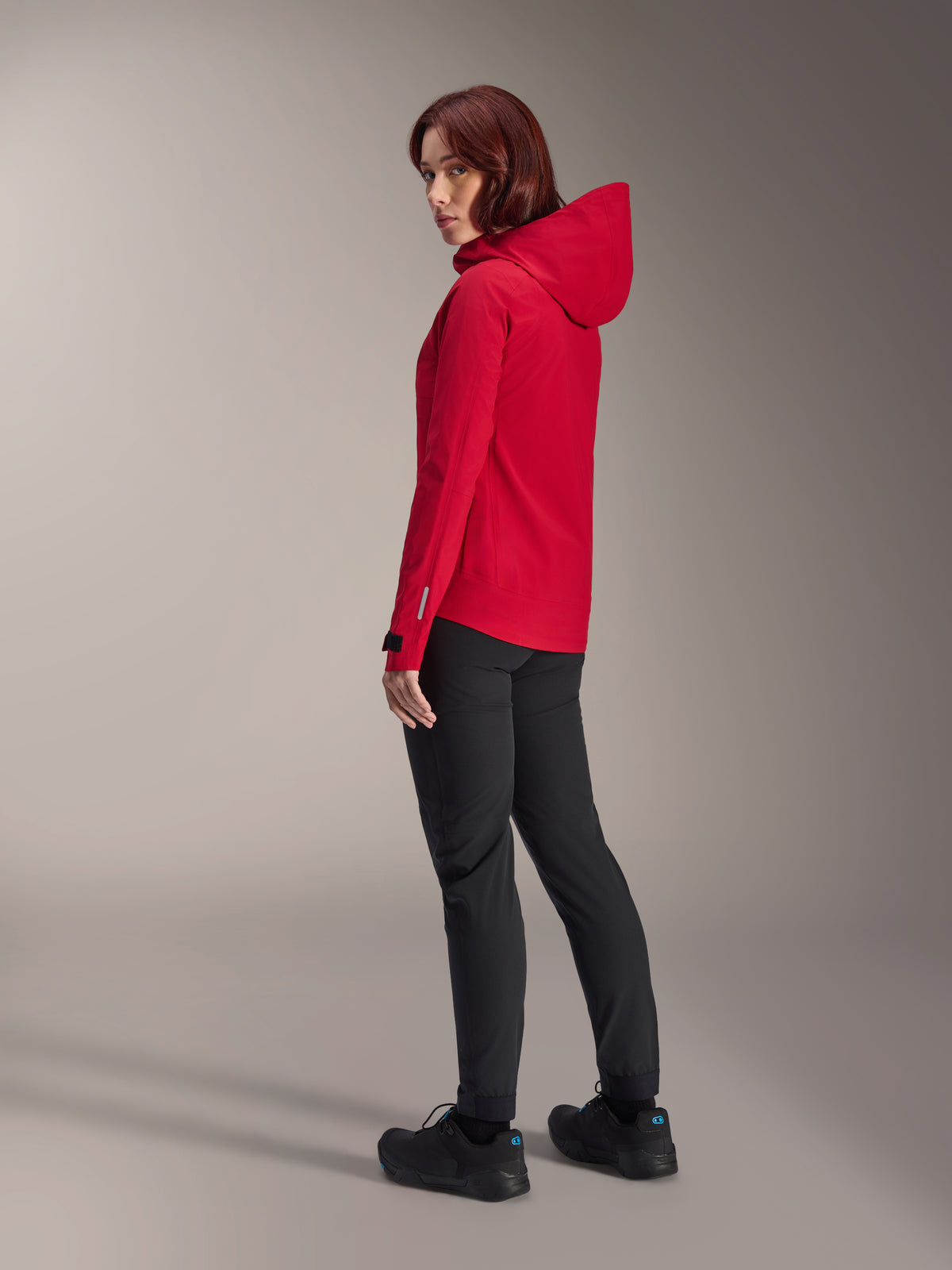 Mulher a usar o casaco impermeável Alpinestars Stella A-Dura Elite em Vermelho Fluo, combinado com calças técnicas de ciclismo pretas e sapatos pretos, vista traseira de três quartos a mostrar o capuz ajustável e a bainha traseira mais longa, vestuário de performance para mountain bike