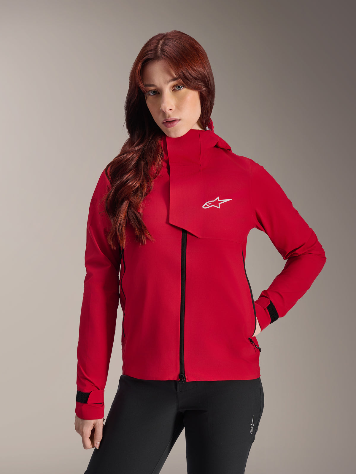 Casaco impermeável Alpinestars Stella A-Dura Elite, casaco de BTT para mulher, Vermelho Fluo, com aba frontal de proteção contra intempéries com logótipo Alpinestars branco, gola alta e punhos ajustáveis, usado com calças pretas para um visual técnico de condução.