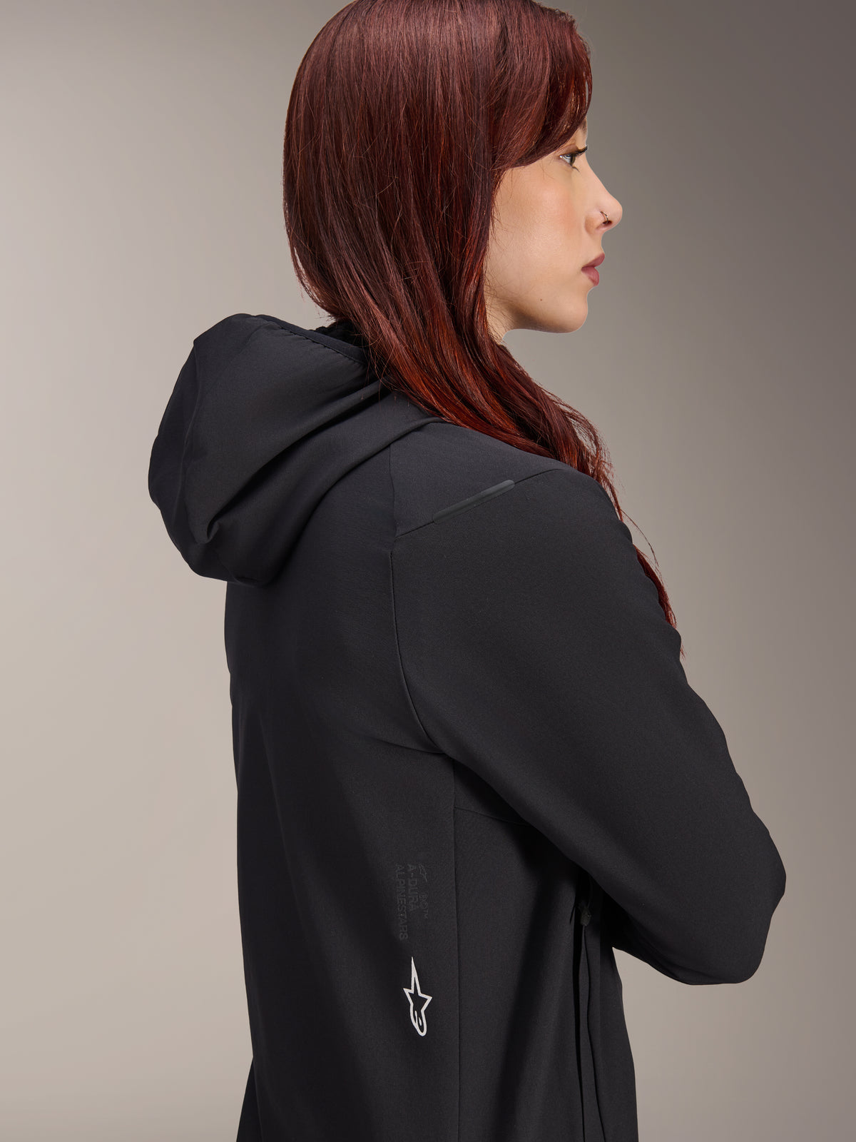 Alpinestars Stella A-Dura Thermal Jacket, casaco de ciclismo feminino, preto, perfil lateral de uma mulher com cabelo ruivo a usar o casaco, apresenta capuz integrado e logótipo da estrela Alpinestars a branco na lateral, equipamento térmico isolado leve para BTT