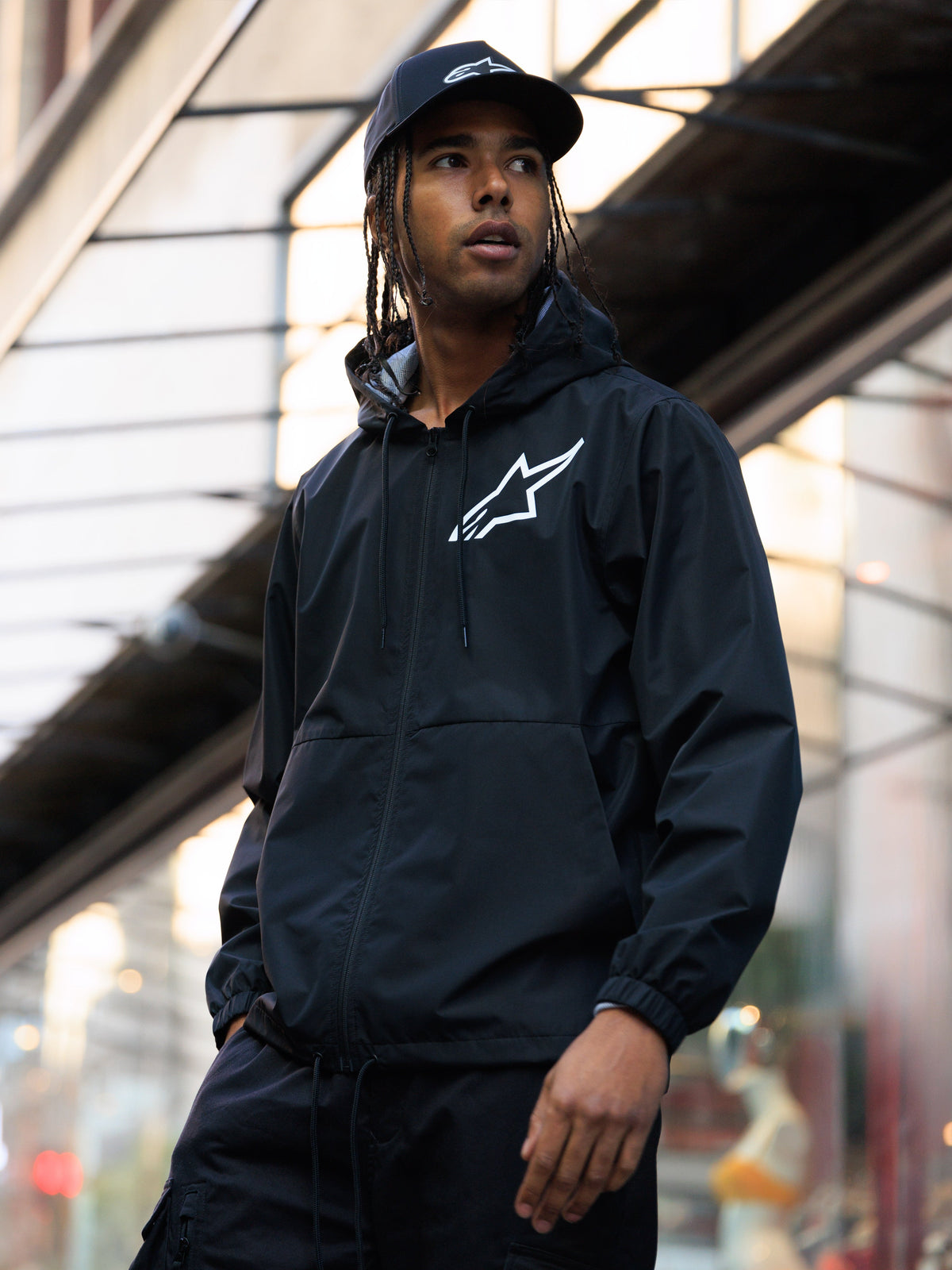 Casaco Corta-Vento Alpinestars Speeder Chromium, preto, corta-vento com capuz e logótipo branco no peito, usado por um homem com boné Alpinestars preto a combinar em ambiente urbano, streetwear casual de motorsport