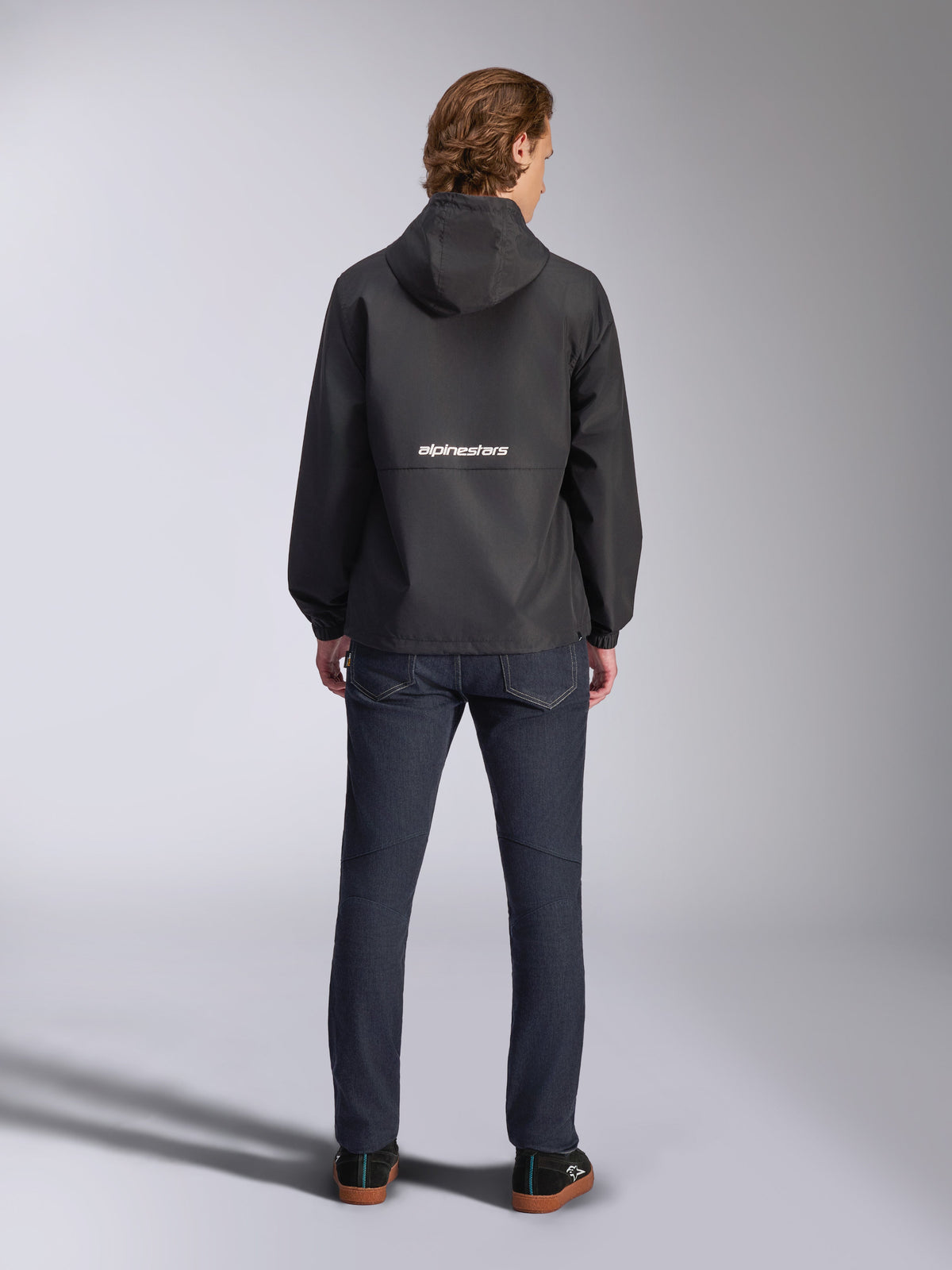 Casaco Corta-Vento Alpinestars Speeder Chromium, preto, vista traseira com logótipo Alpinestars branco ao centro, estilo com capuz, usado por modelo com calças de ganga escuras, casaco casual inspirado no desporto motorizado