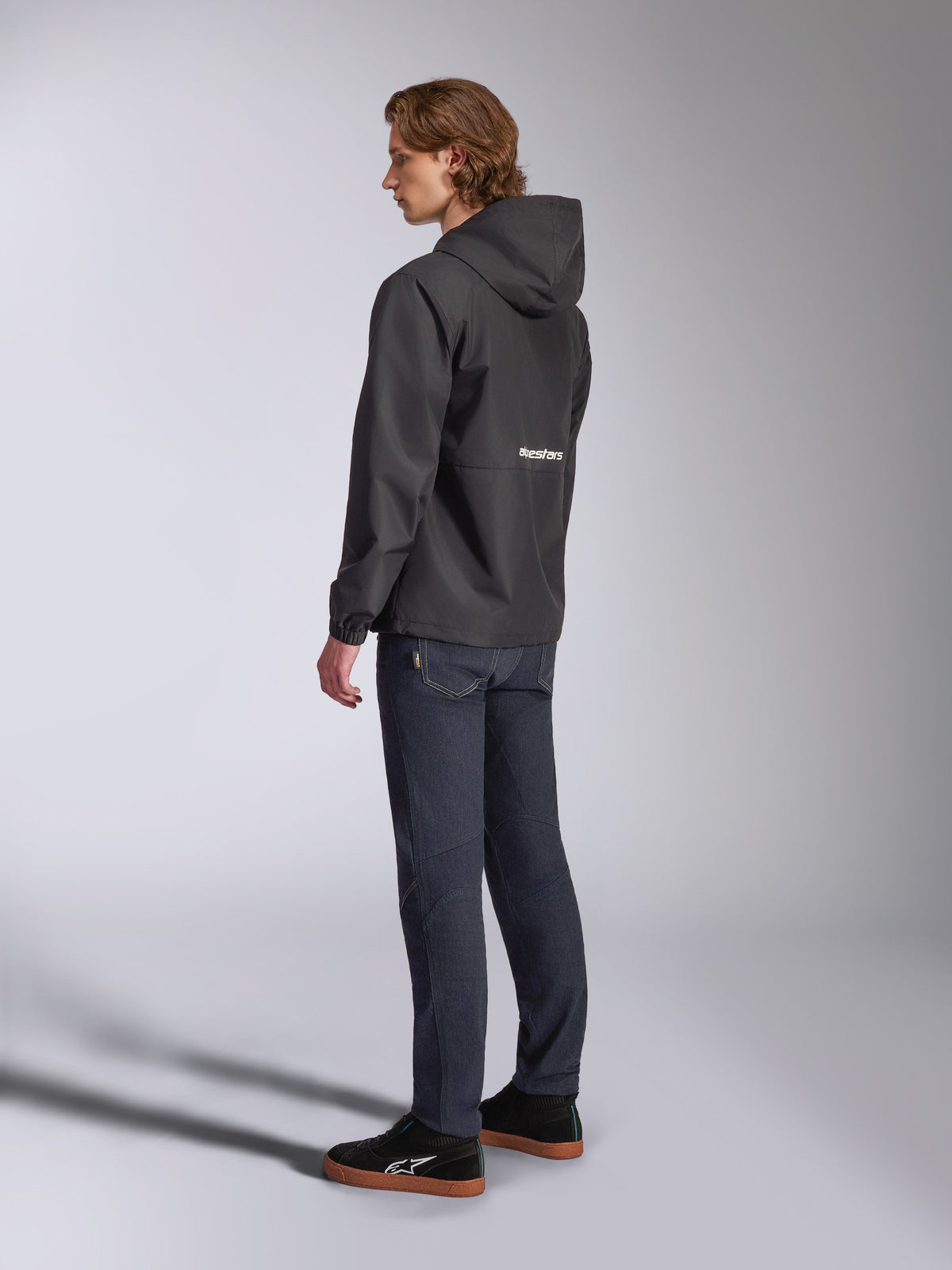 Casaco Corta-Vento Alpinestars Speeder Chromium, preto com logótipo branco nas costas, corta-vento casual para homem com capuz, vista de perfil lateral usado com calças de ganga escuras e sapatilhas pretas, streetwear inspirado no desporto motorizado