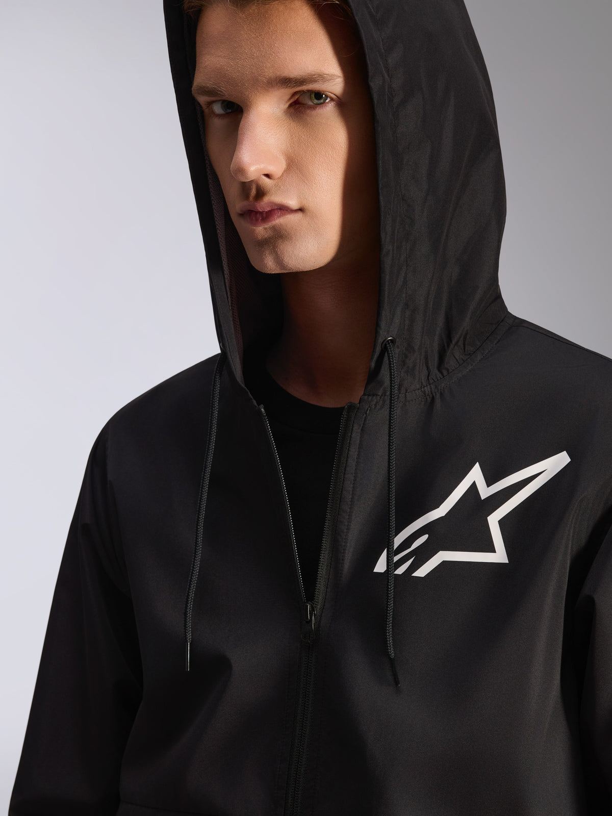 Alpinestars Speeder Chromium Windbreak Jacket, preto, grande plano de um corta-vento com capuz e logótipo de estrela branca no peito, inclui capuz com cordão, material têxtil leve, streetwear casual inspirado no desporto motorizado