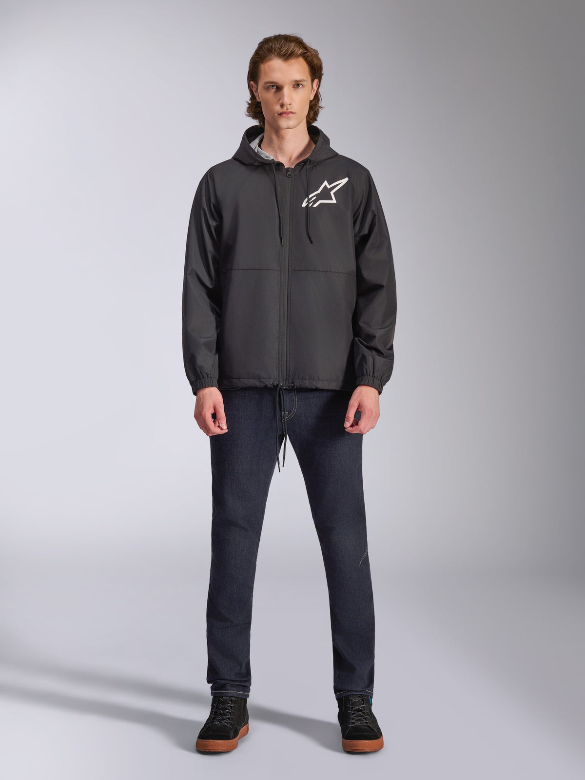 Casaco Corta-Vento Alpinestars Speeder Chromium, preto com logótipo Astars branco, vestido por modelo com calças de ganga azul-escuro e sapatilhas pretas, com design com capuz e fecho frontal, streetwear casual inspirado no desporto motorizado