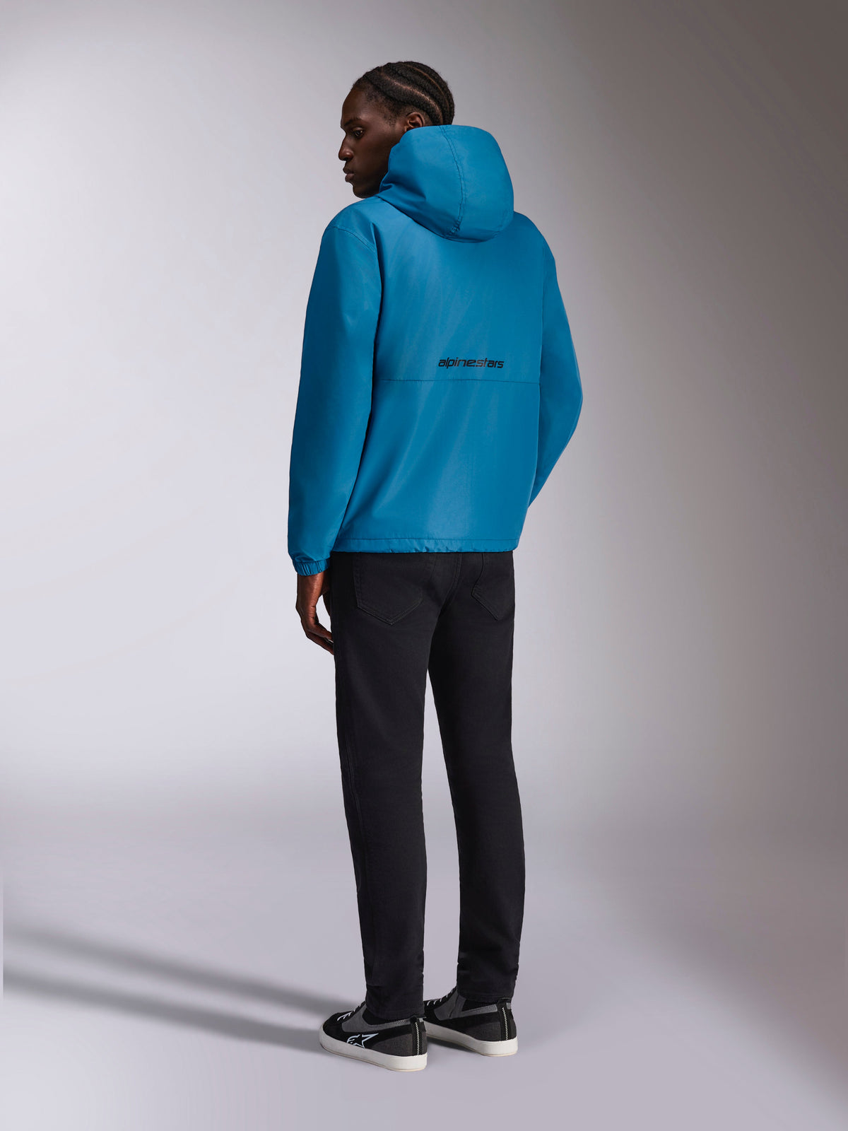 Casaco Corta-Vento Alpinestars Speeder Chromium, corta-vento com capuz, azul, vista traseira com logótipo Alpinestars preto nas costas, usado com calças pretas e sapatilhas pretas e brancas, streetwear casual inspirado no desporto motorizado