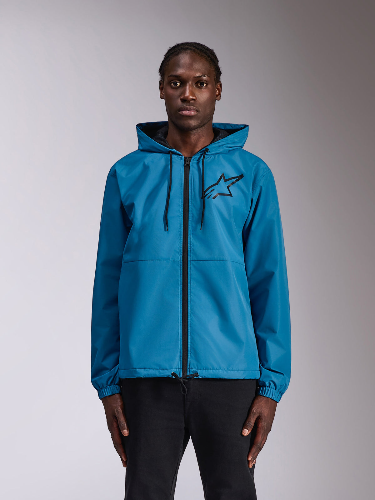 Casaco Corta-Vento Alpinestars Speeder Chromium, corta-vento azul com capuz, logótipo Astars preto no peito, fecho de correr frontal, punhos elásticos e capuz com cordões, usado por um modelo como vestuário desportivo casual.