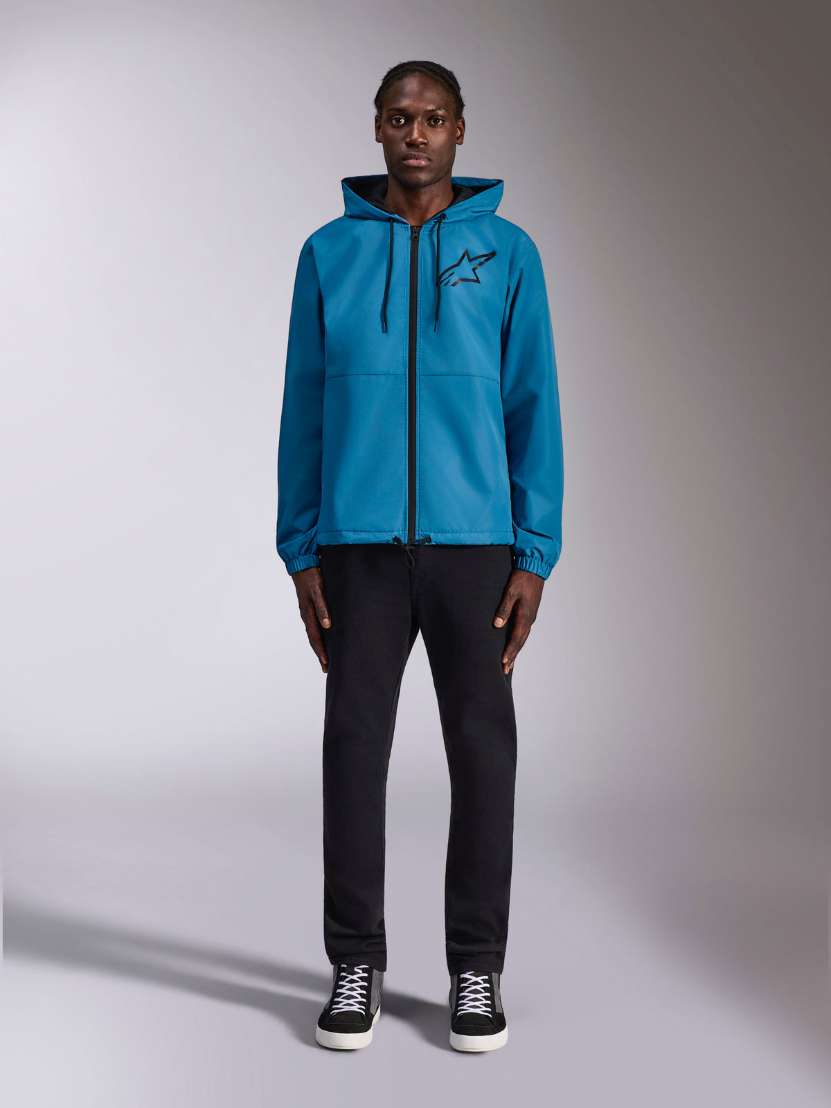 Modelo a usar o Casaco Corta-vento Alpinestars Speeder Chromium em azul, com design com capuz e cordões pretos, fecho de correr frontal preto e logótipo Astars preto no peito, combinado com calças e sapatilhas pretas, streetwear casual de desporto motorizado