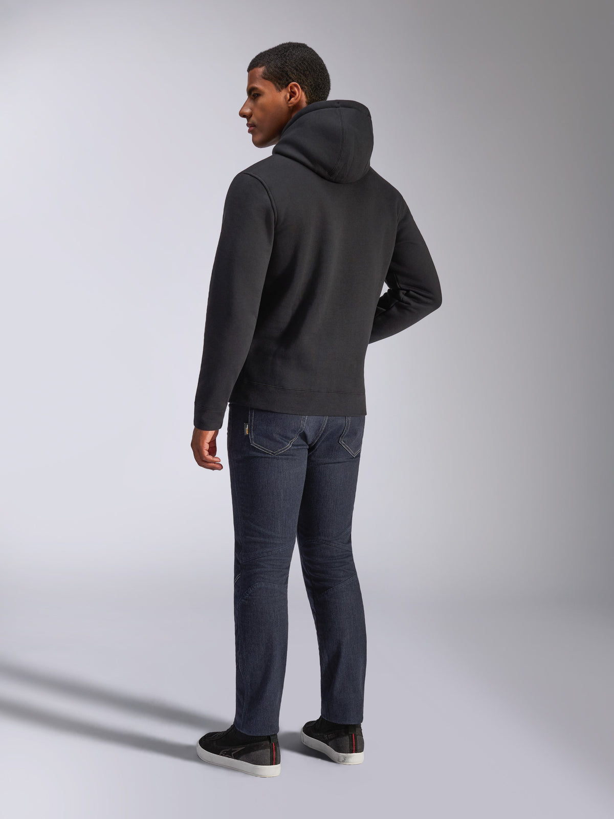 Alpinestars Blaze V3 Hoodie, vista traseira de um homem a usar a camisola preta com capuz, combinada com calças de ganga escuras e sapatilhas pretas, streetwear casual inspirado no desporto motorizado