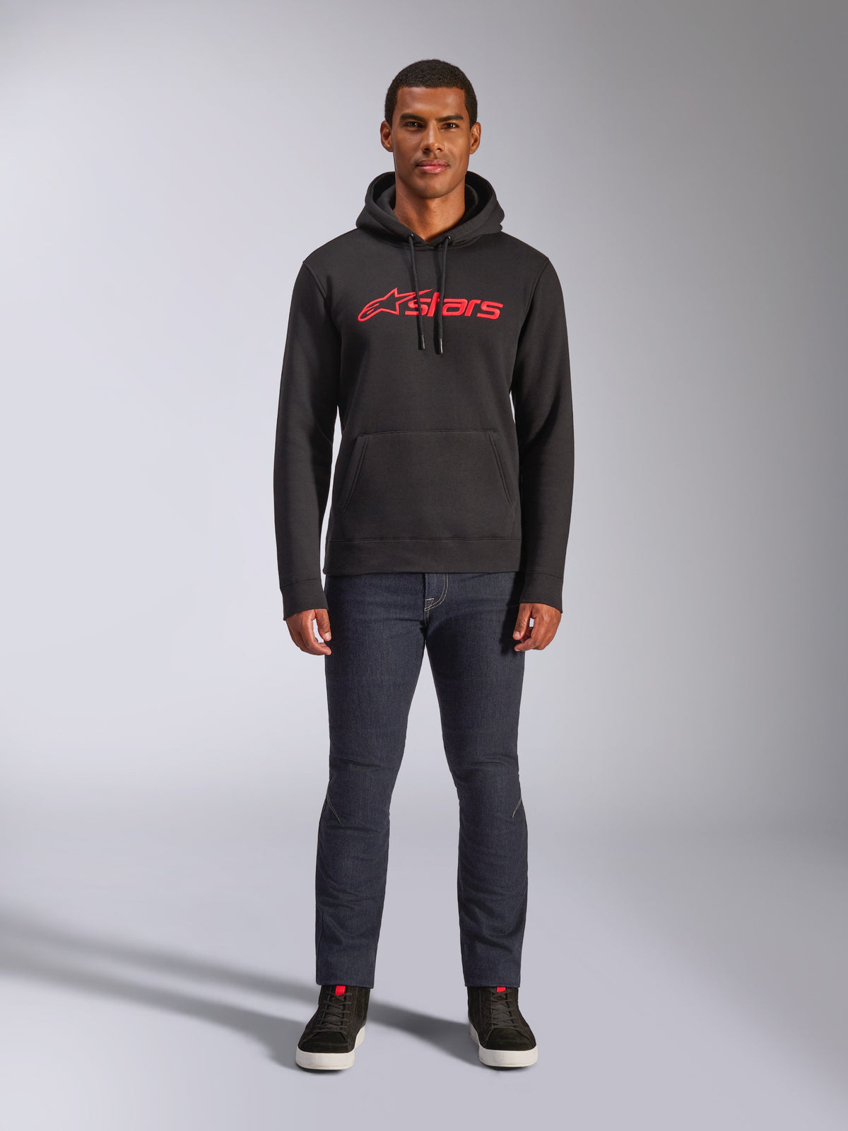 Alpinestars Blaze V3 Hoodie, camisola com capuz de estilo streetwear casual, preta com logótipo Astars vermelho, vista frontal, usada por um modelo com calças de ganga escuras e sapatilhas pretas, design moderno inspirado no desporto motorizado
