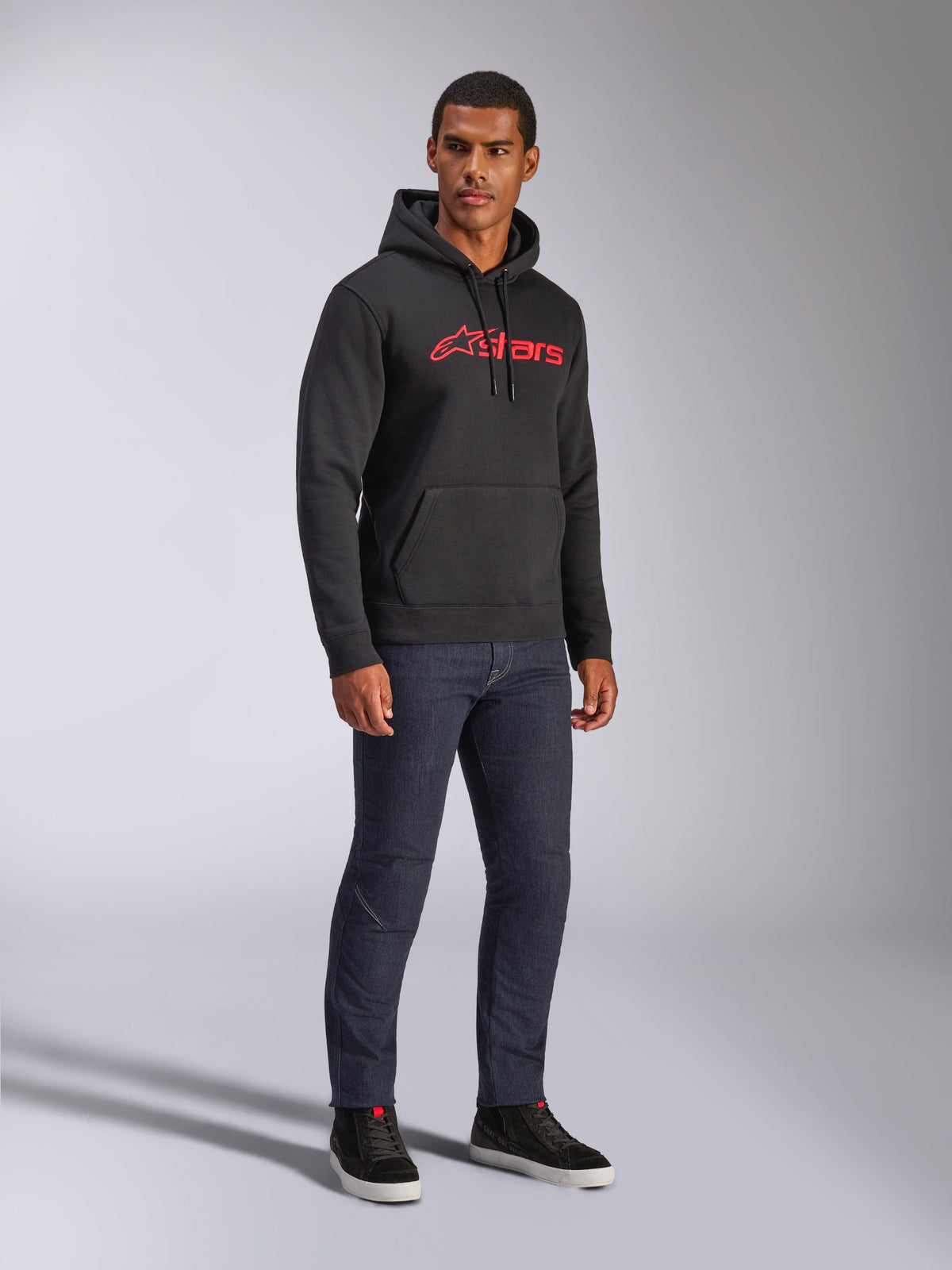 Alpinestars Blaze V3 Hoodie, camisola com capuz preta com logótipo Alpinestars vermelho no peito, usada por um modelo masculino com calças de ganga escuras e sapatilhas pretas, streetwear casual inspirado no desporto motorizado