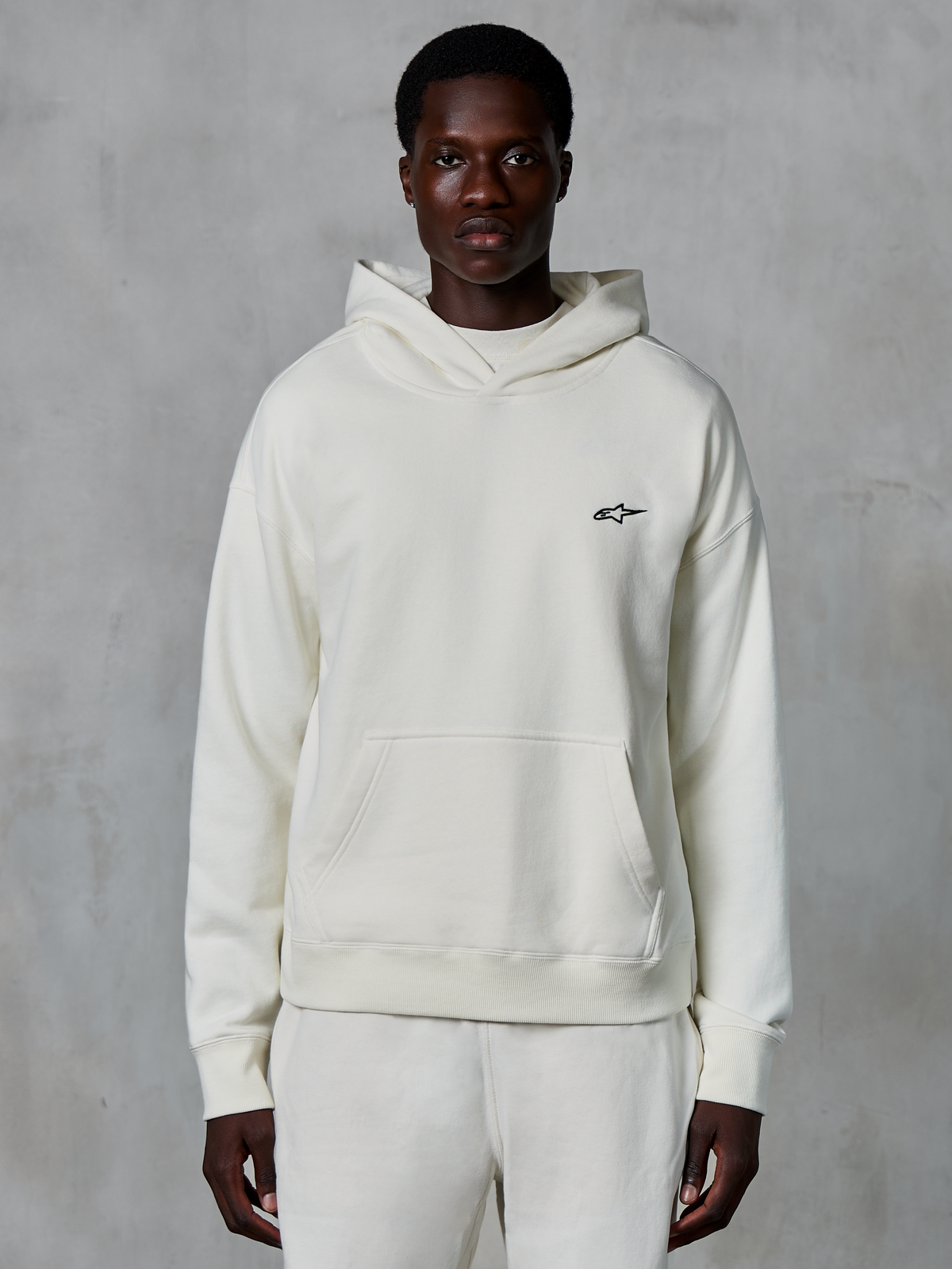 Hoodie Alpinestars Essentials Ageless, off-white, usado por modelo masculino, logótipo pequeno preto no peito, bolso frontal tipo canguru, mangas compridas, streetwear casual inspirado no desporto motorizado
