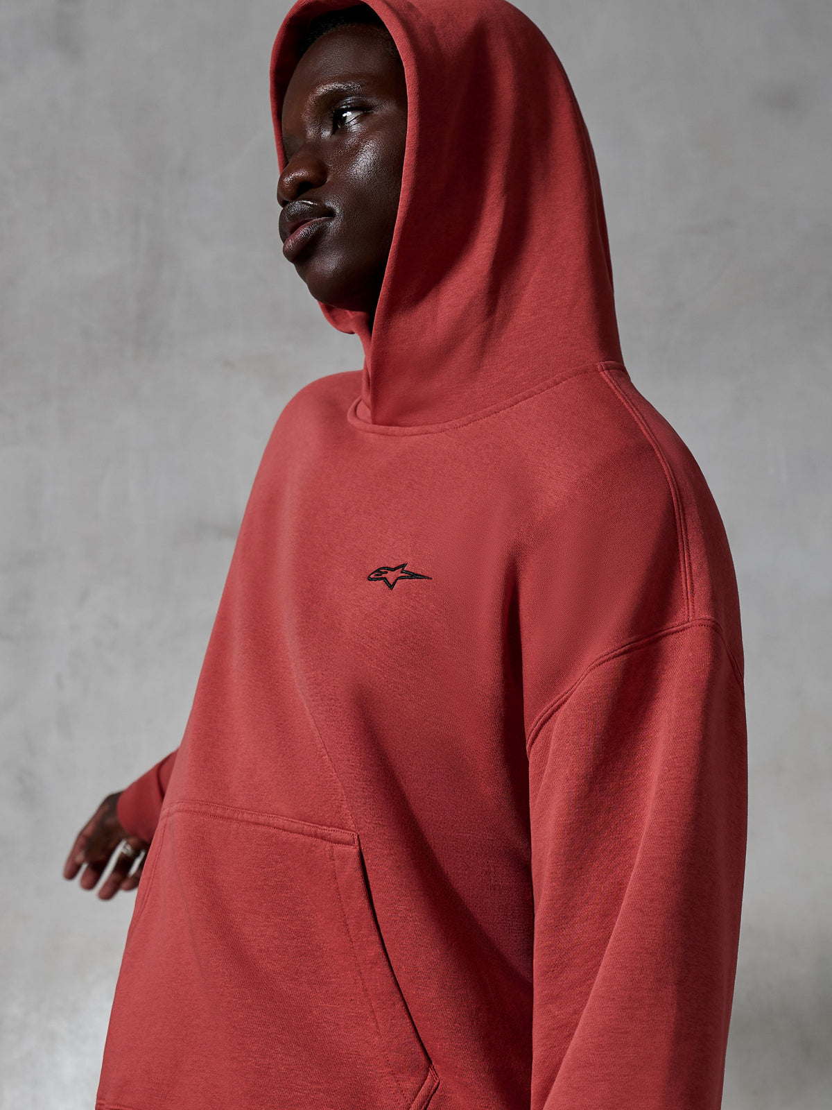 Alpinestars Essentials Ageless Hoodie, roupa desportiva casual, vermelho tijolo, usado por um modelo com o capuz levantado, com um pequeno logótipo preto bordado, textura de fleece macio, estilo streetwear de ajuste descontraído