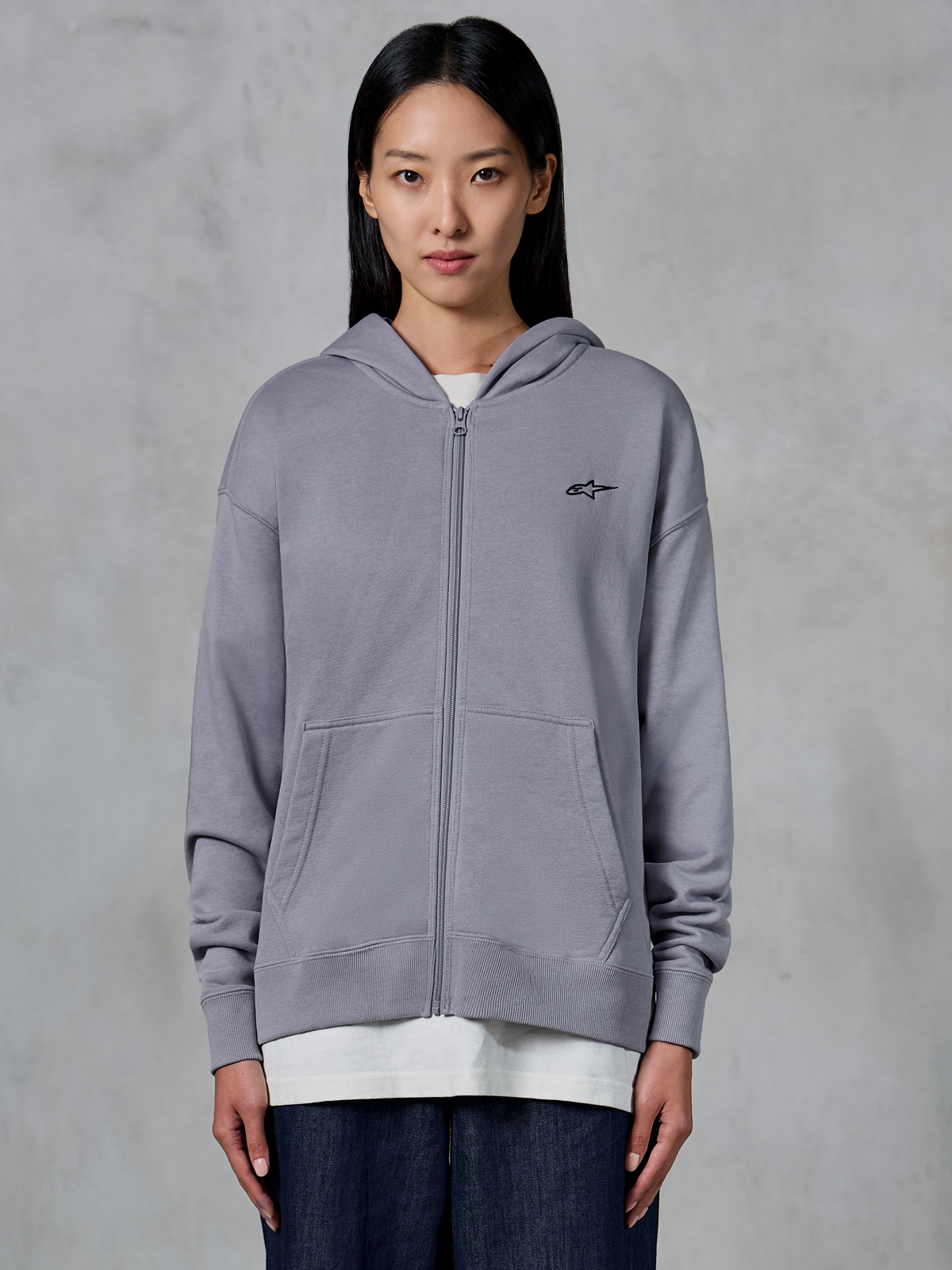 Alpinestars Essentials Ageless Zip Hoodie, prateado, streetwear casual, sweatshirt de fleece com fecho integral e pequeno logótipo Alpinestars preto no peito, bolso canguru dividido, usado por modelo sobre t-shirt branca e jeans escuros