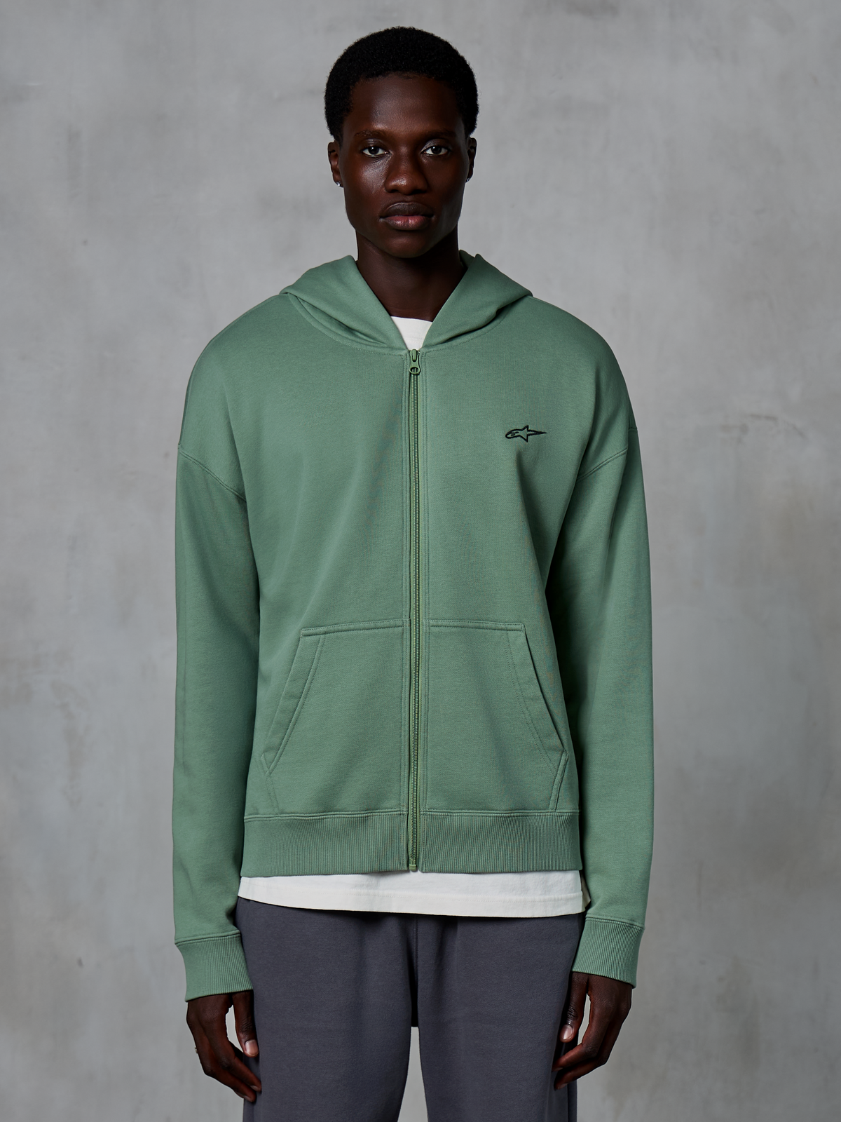 Alpinestars Essentials Ageless Zip Hoodie, hoodie de streetwear casual com fecho, verde Cactus, com um logótipo bordado subtil no peito, bolsos tipo canguru divididos e punhos canelados, vestido por um modelo masculino num fundo cinzento texturizado.
