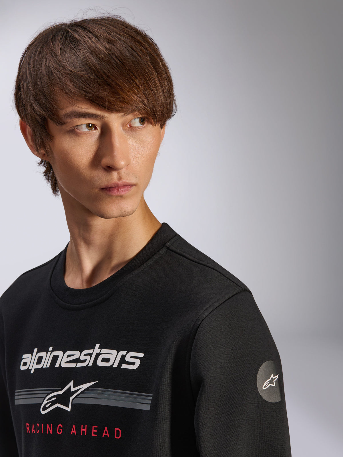 Sweatshirt Alpinestars Bettern Crew, preta, com logótipo Alpinestars branco, riscas cinzentas e texto 'RACING AHEAD' a vermelho, logótipo circular com estrela na manga, streetwear casual inspirado no desporto motorizado