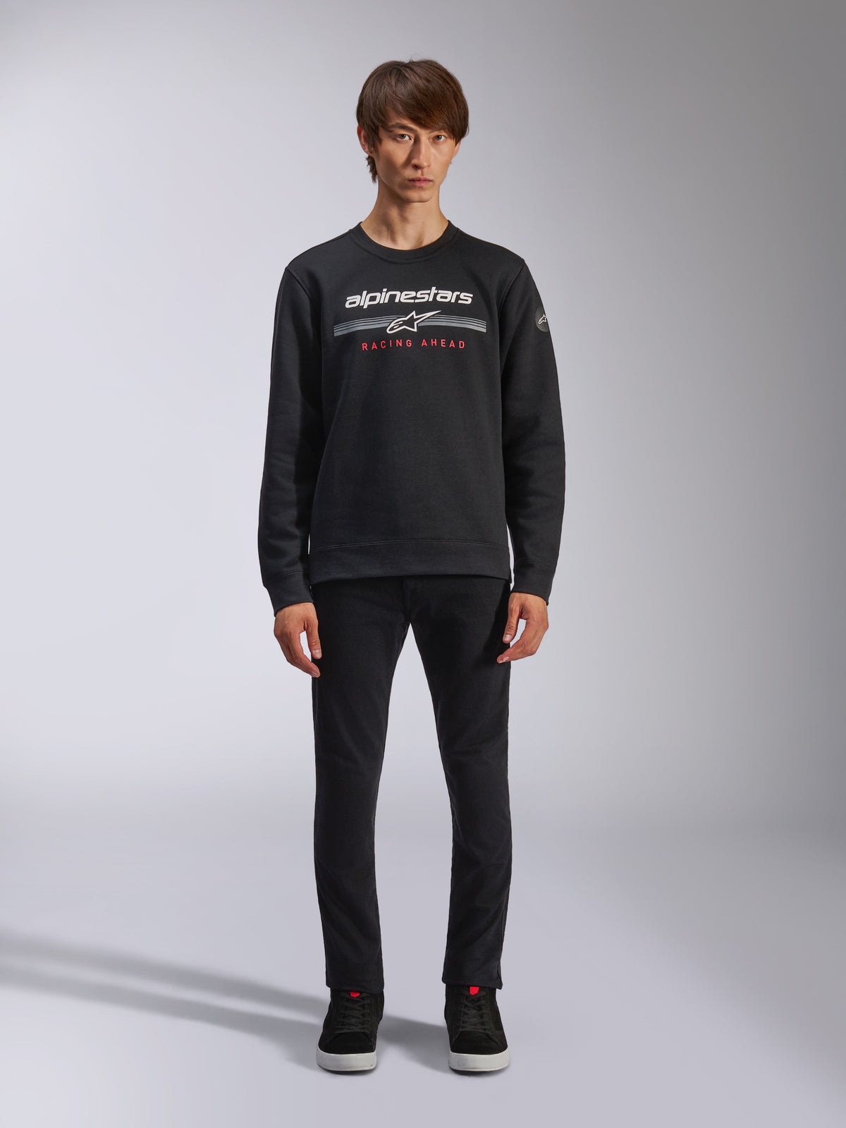 Alpinestars Bettern Crew, camisola de gola redonda preta, apresenta o logótipo Alpinestars em branco com riscas horizontais cinzentas e o texto 'RACING AHEAD' em vermelho, streetwear casual de desportos motorizados, vestida por um modelo com calças pretas e sapatilhas