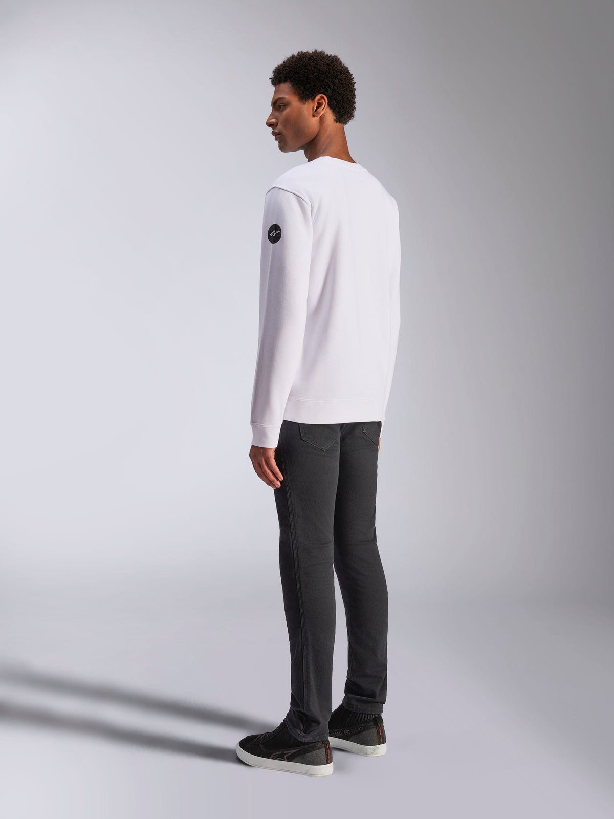 Alpinestars Bettern Crew, sweatshirt branca de gola redonda, vista lateral e traseira, logótipo circular preto na manga, usada com calças cinzentas escuras e ténis pretos, streetwear casual de estilo de vida motorsport