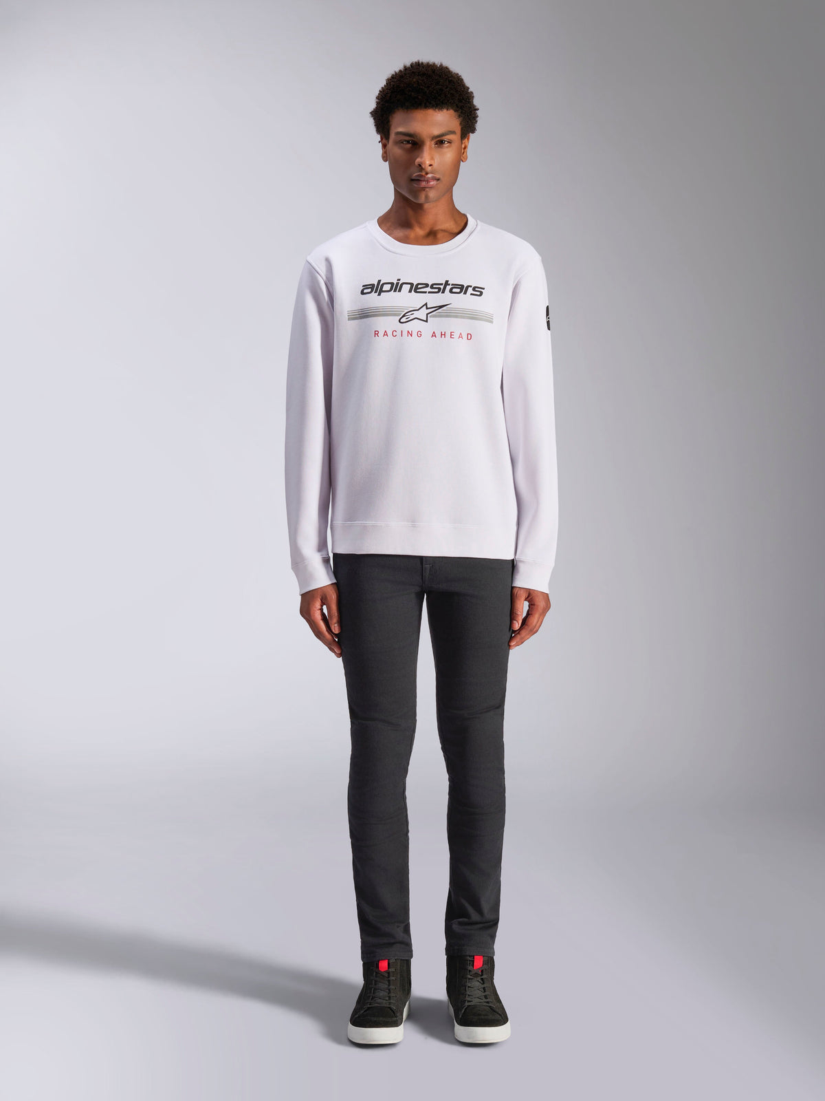 Alpinestars Bettern Crew, sweatshirt branca de gola redonda, usada por um modelo masculino com calças cinzento-escuras e sapatilhas de cano alto pretas, apresentando a marca Alpinestars com o texto vermelho 'Racing Ahead', streetwear casual inspirado no desporto motorizado