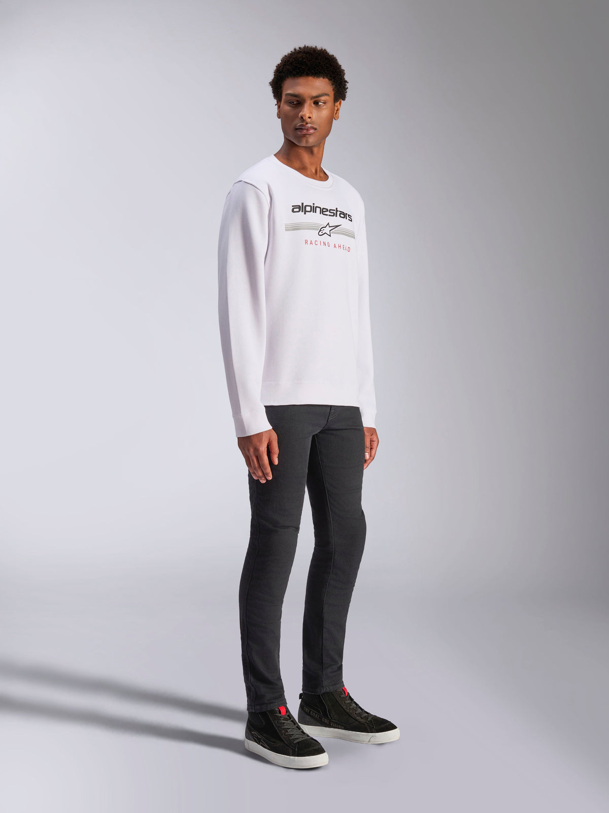 Alpinestars Bettern Crew, sweatshirt pullover branca, com o logótipo Alpinestars em preto e o texto "Racing Ahead" em vermelho, streetwear casual de desportos motorizados, usada por um modelo com calças escuras e sapatilhas pretas