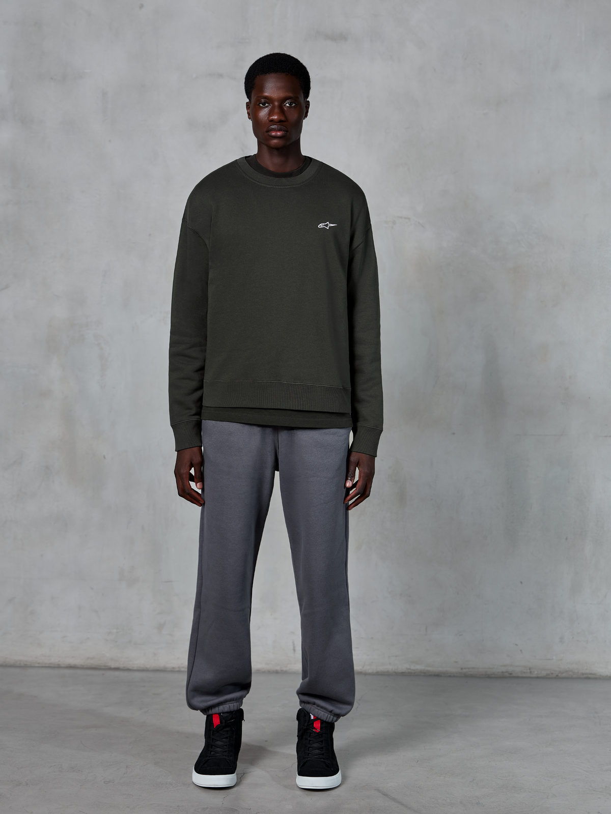Alpinestars Essentials Ageless Crew, sweatshirt de gola redonda verde-escuro com um pequeno logótipo branco no peito, usada por um modelo com calças de fato de treino cinzentas e sapatilhas de cano alto pretas, vestuário casual lifestyle