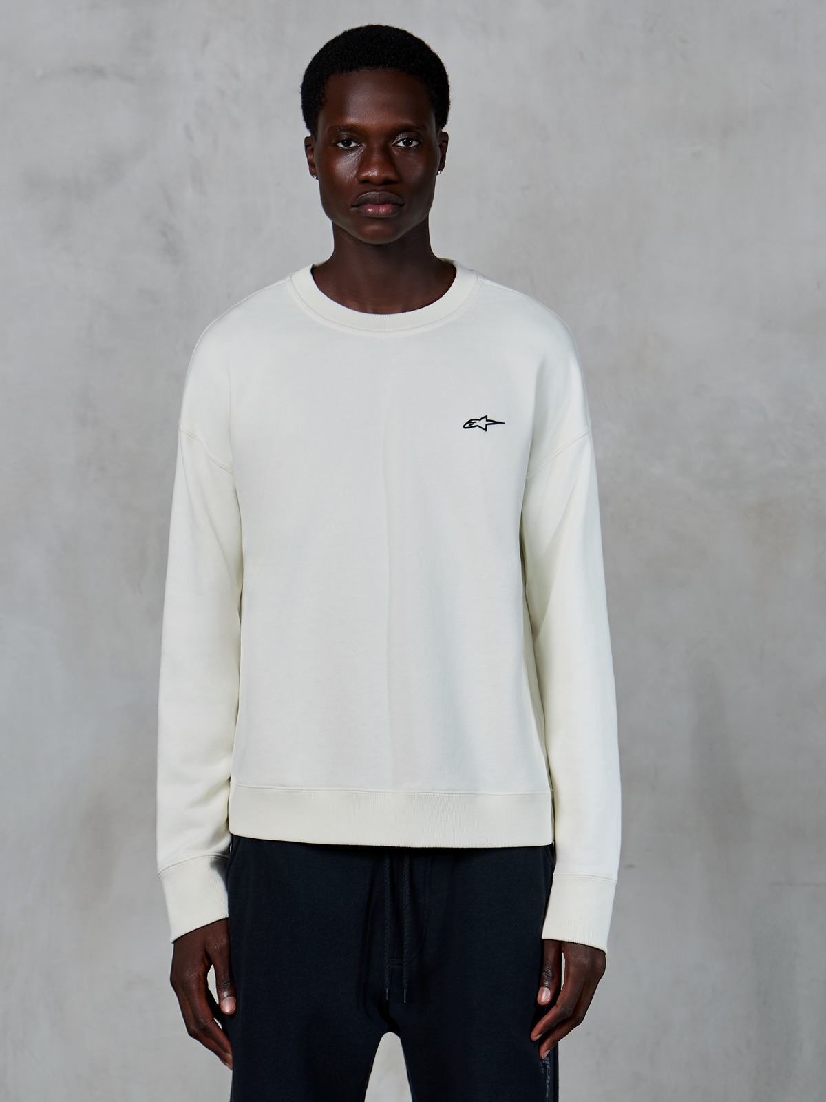 Alpinestars Essentials Ageless Crew, sweatshirt de gola redonda off-white, pequeno logótipo Astars preto no lado esquerdo do peito, vestuário desportivo casual, textura de fleece premium, usada por modelo com calças joggers pretas
