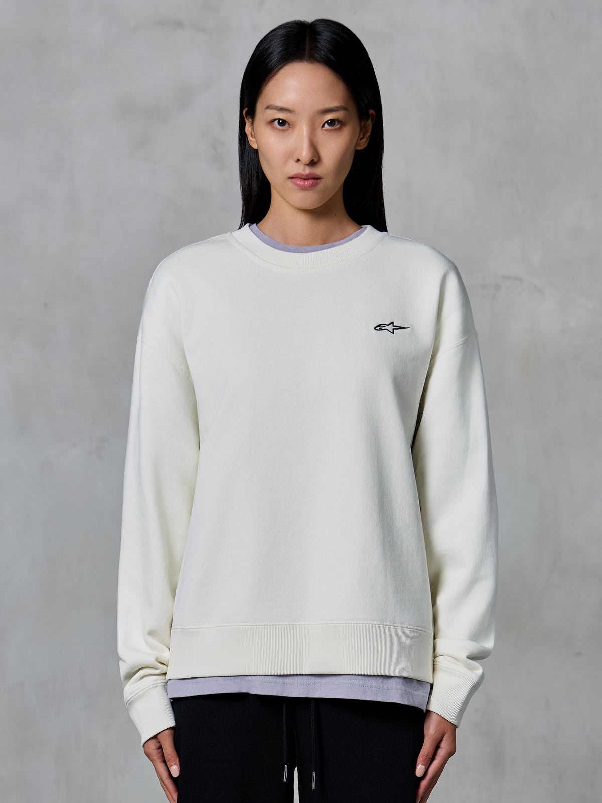 Alpinestars Essentials Ageless Crew, sweatshirt de gola redonda, off white, com um pequeno logótipo preto da Alpinestars no peito, vestuário de estilo de vida casual modelado numa mulher